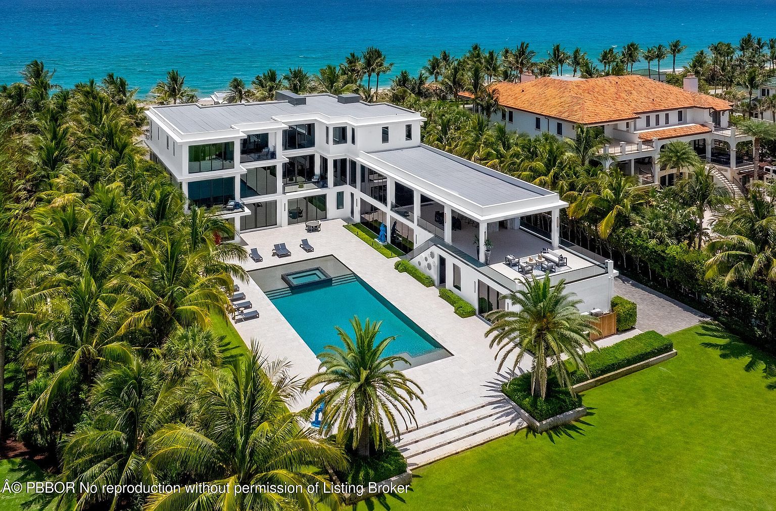 This aerial view showcases a luxurious modern home with a large pool and spa, surrounded by lush tropical landscaping and palm trees. The property features a sleek, white exterior with expansive windows and a flat roof, offering stunning ocean views. The overall impression is one of high-end coastal living.