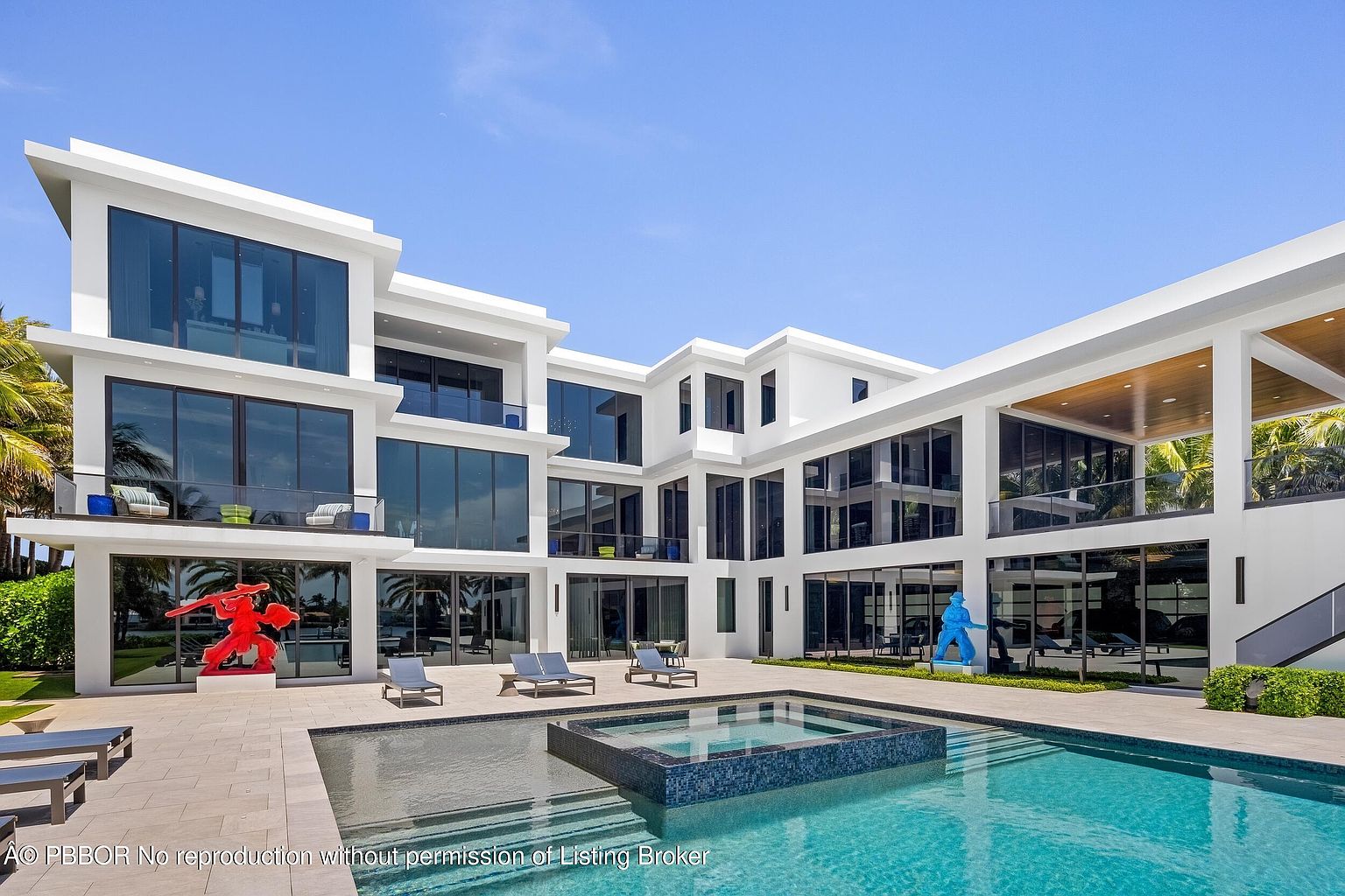 This image showcases the rear exterior of a modern luxury home, emphasizing the pool and spa area. The architecture features clean lines, large windows, and a white facade. The pool area includes lounge chairs, sculptures, and a built-in spa, creating an inviting and upscale outdoor living space.
