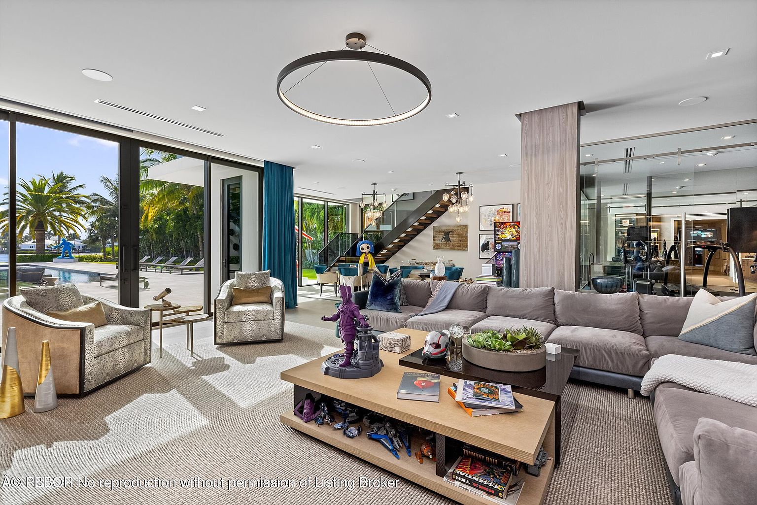 This is a spacious and modern living room featuring a large sectional sofa, a unique coffee table with display shelves, and floor-to-ceiling windows that offer views of the outdoor pool area. The room is well-lit with a circular pendant light and natural light, creating a bright and inviting atmosphere. A staircase is visible in the background, adding architectural interest.