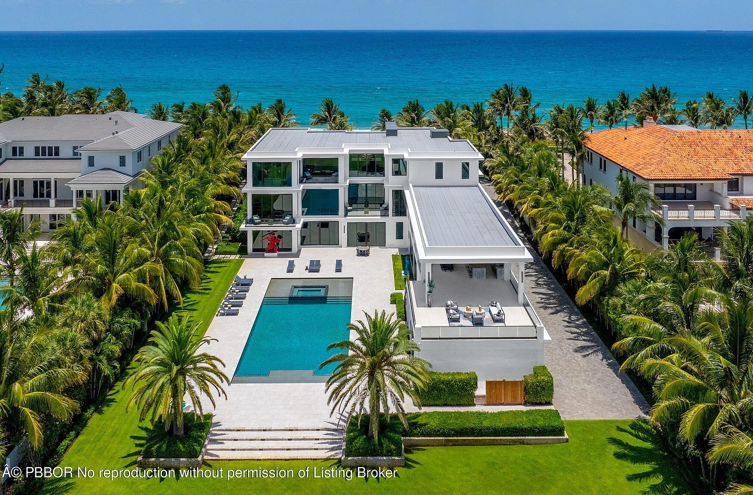This aerial view showcases a luxurious modern home with a pool and meticulously landscaped grounds. The property features a multi-story structure with expansive windows, a flat roof, and a spacious outdoor patio area. The turquoise ocean backdrop and surrounding palm trees enhance the property's appeal, creating a serene and upscale atmosphere.