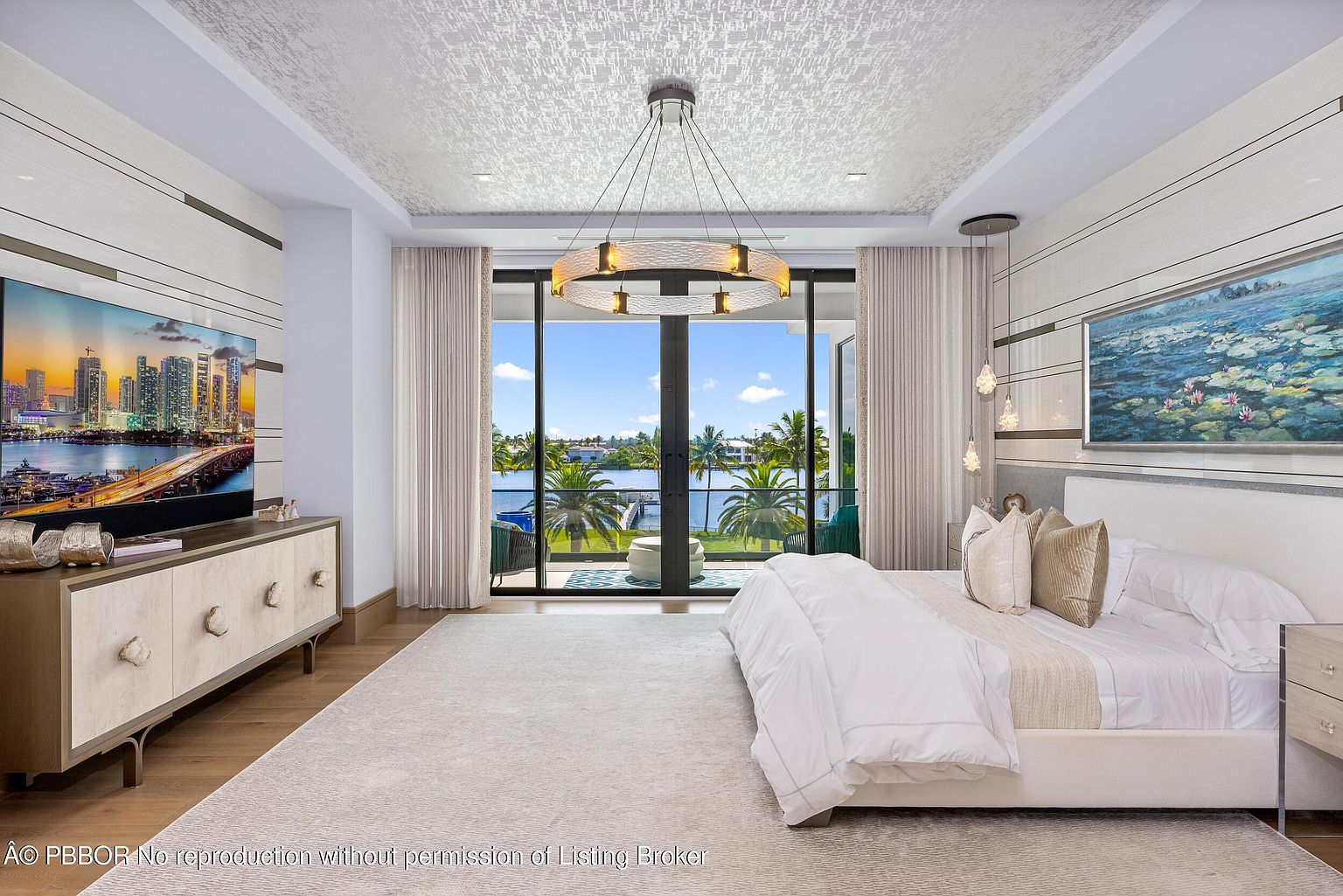 This is a luxurious primary bedroom featuring a king-size bed with white linens and decorative pillows, a modern chandelier, and floor-to-ceiling windows offering a stunning waterfront view. The room is decorated in neutral tones with elegant wall paneling and artwork, creating a serene and sophisticated atmosphere. A large area rug covers the hardwood floor, adding warmth and texture to the space.