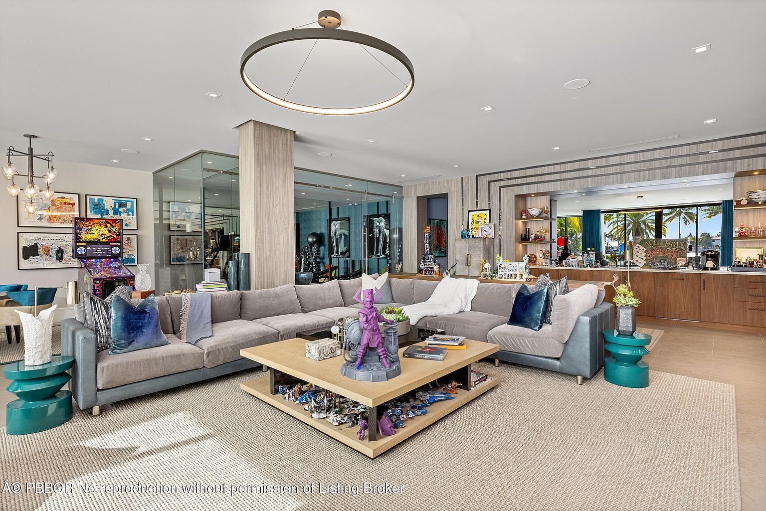 This is a spacious and luxurious living room featuring a large sectional sofa, a unique coffee table with display shelves, and a modern circular light fixture. The room is well-lit and decorated with various art pieces and decorative items, creating a sophisticated and inviting atmosphere. A pinball machine is visible in the background, adding a playful touch to the space.