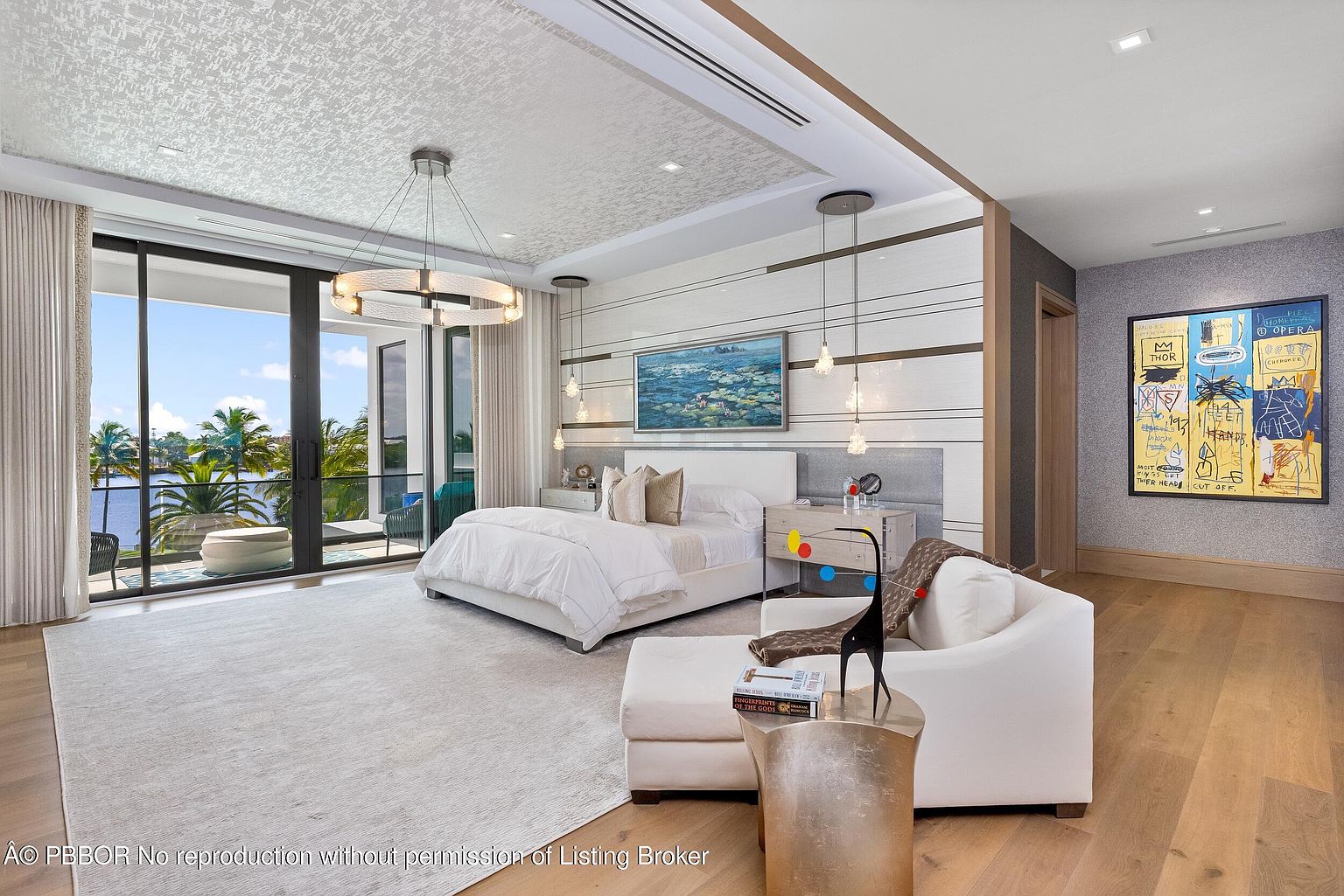 This is a luxurious primary bedroom featuring a king-size bed with white linens, a modern chandelier, and floor-to-ceiling windows offering a view of the water. The room is decorated in neutral tones with a statement art piece, creating a serene and sophisticated atmosphere. A seating area with a white sofa and a unique side table adds to the room's elegance and functionality.