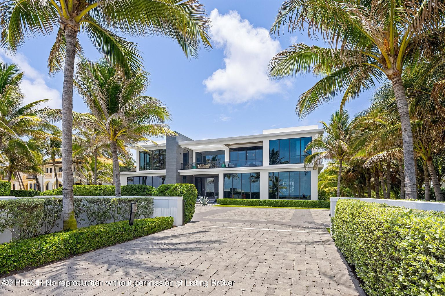 This is a front exterior view of a modern, two-story home framed by lush palm trees and manicured hedges. The house features large windows and a balcony, suggesting ample natural light and outdoor living space. A brick driveway leads to the entrance, enhancing the property's curb appeal and upscale feel.