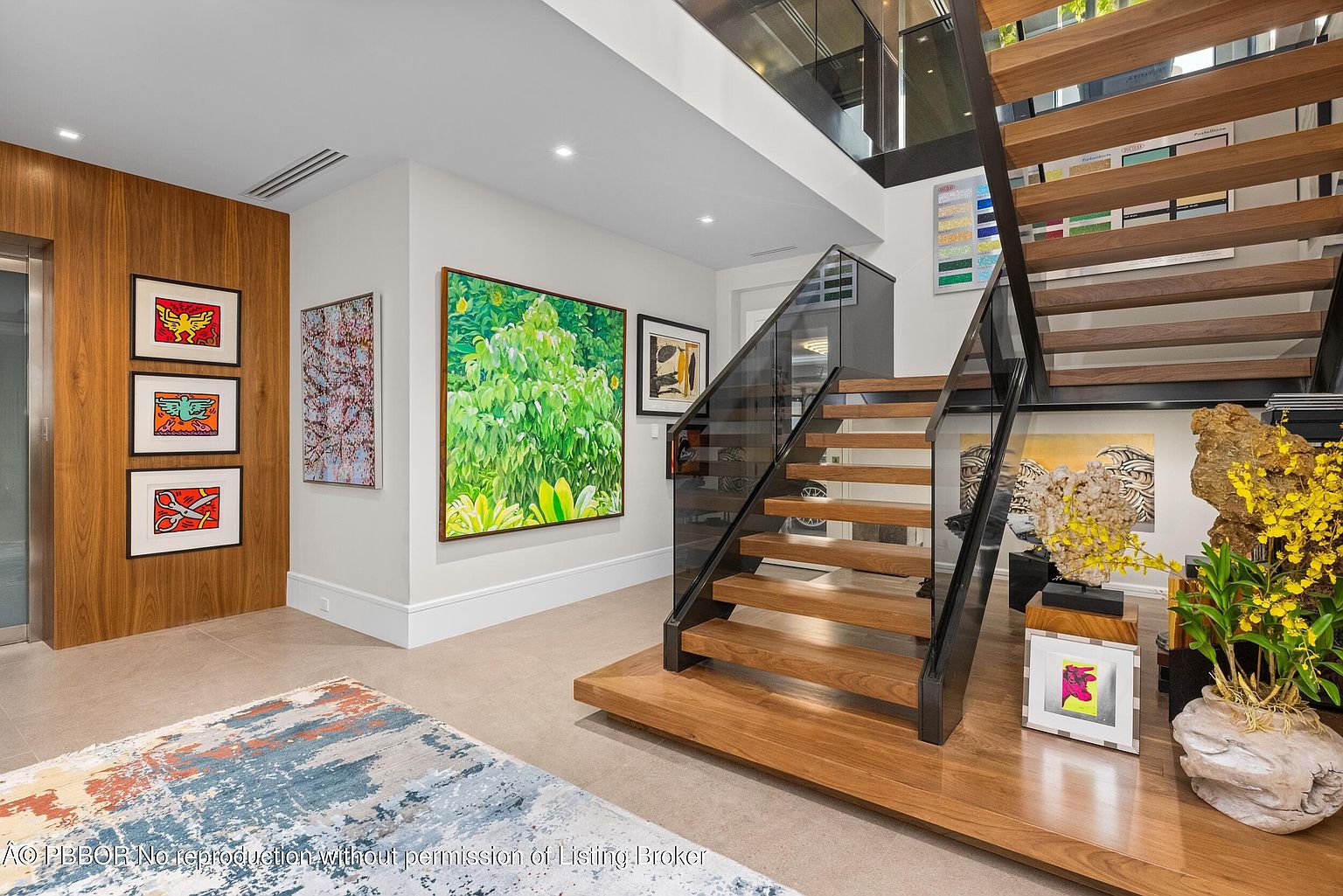 This interior shot showcases a modern hallway with a striking staircase featuring wooden steps and glass railings. Artwork adorns the walls, adding a touch of sophistication and color to the space. The flooring is a neutral tile, and the overall impression is one of contemporary elegance and artistic flair.