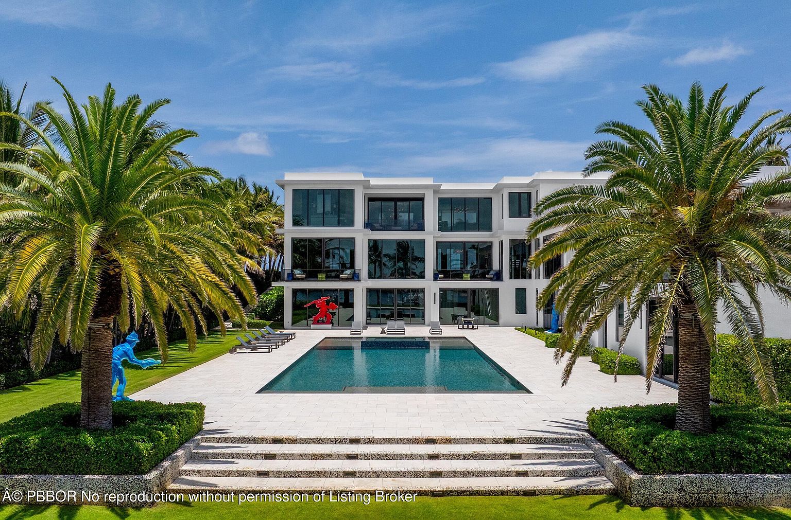 This image showcases the rear exterior of a modern, multi-story home, emphasizing the pool and outdoor living space. The pool is surrounded by a light-colored patio, with palm trees framing the view and adding a tropical feel. Sculptures are placed on the lawn, adding an artistic touch to the property.