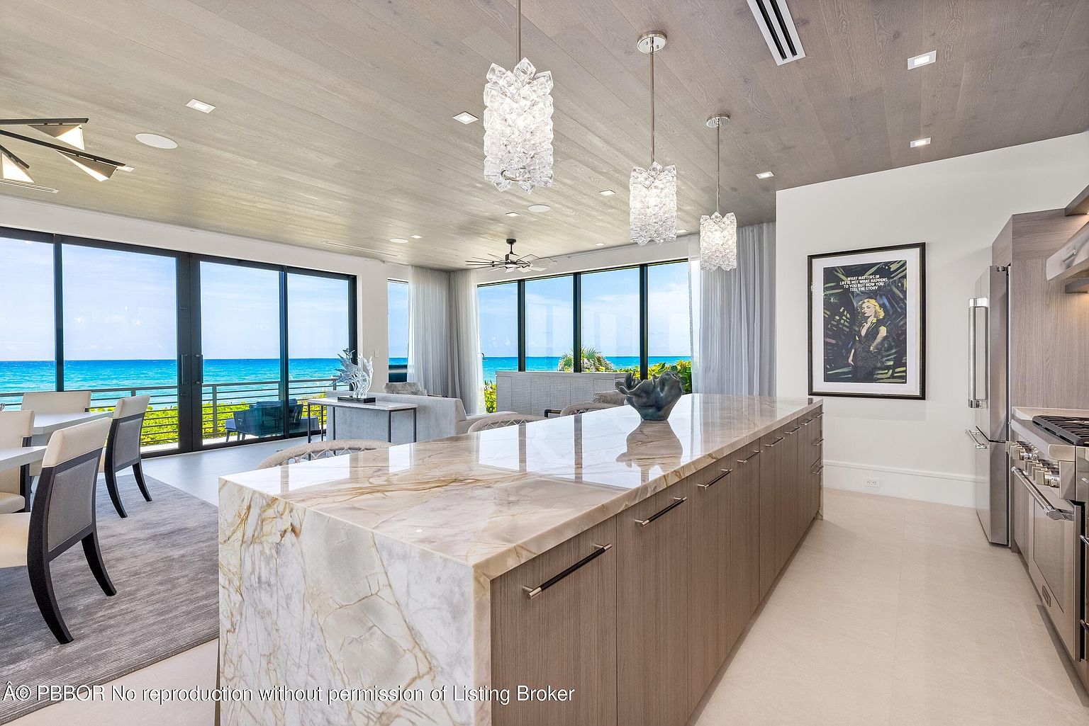 This interior shot showcases a modern kitchen with a large island featuring a marble countertop and wooden cabinetry. The kitchen opens to a living area with floor-to-ceiling windows offering stunning ocean views. Three decorative pendant lights hang above the island, adding a touch of elegance.