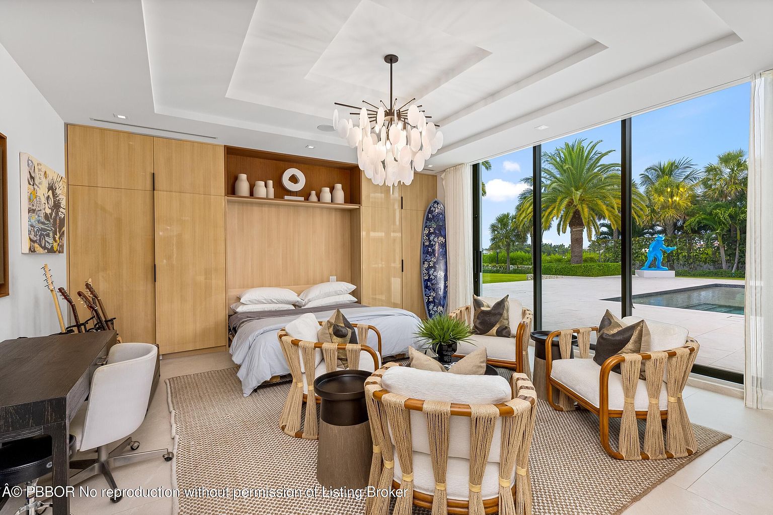 This primary bedroom features a built-in bed with light wood cabinetry and shelving, complemented by a unique chandelier. The room includes a seating area with stylish rope-accented chairs and a large window offering views of the pool and landscaped yard. The overall design creates a serene and luxurious atmosphere.