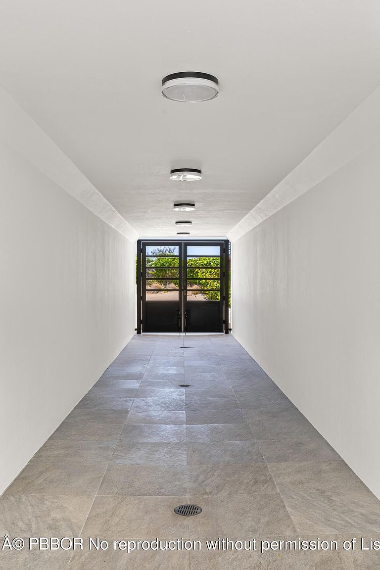 This is an interior shot of a long hallway with gray tiled flooring and white walls. The hallway is illuminated by several ceiling-mounted lights, leading to a set of black metal doors at the end, which provide a view of the outdoors. The perspective emphasizes the length of the hallway, creating a sense of depth and spaciousness.