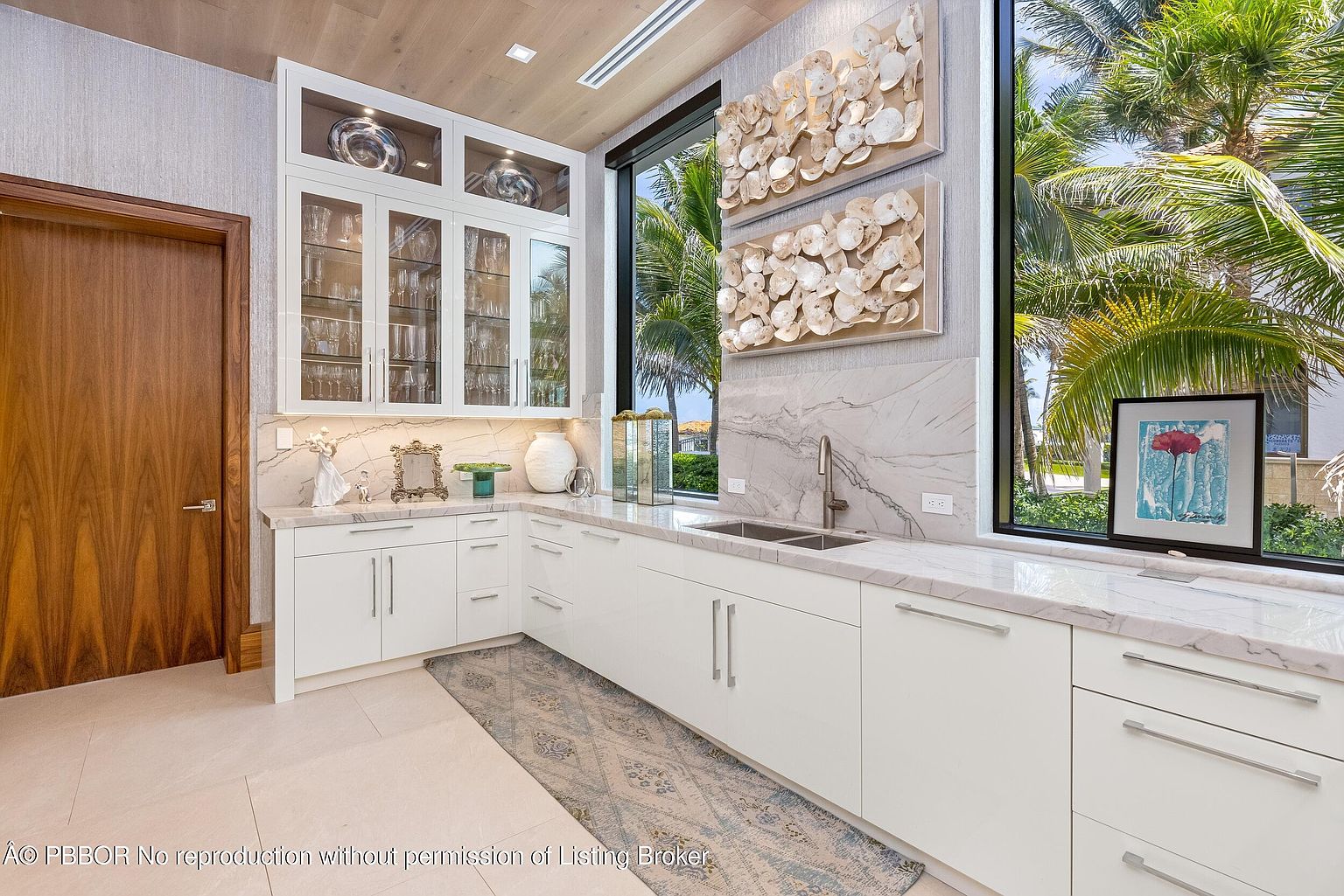 This interior shot showcases a modern kitchen with white cabinetry, marble countertops, and a built-in glass-front display cabinet. A large window provides natural light and a view of palm trees, while decorative shell art adorns the wall. The space is clean, bright, and designed with a sophisticated coastal aesthetic.