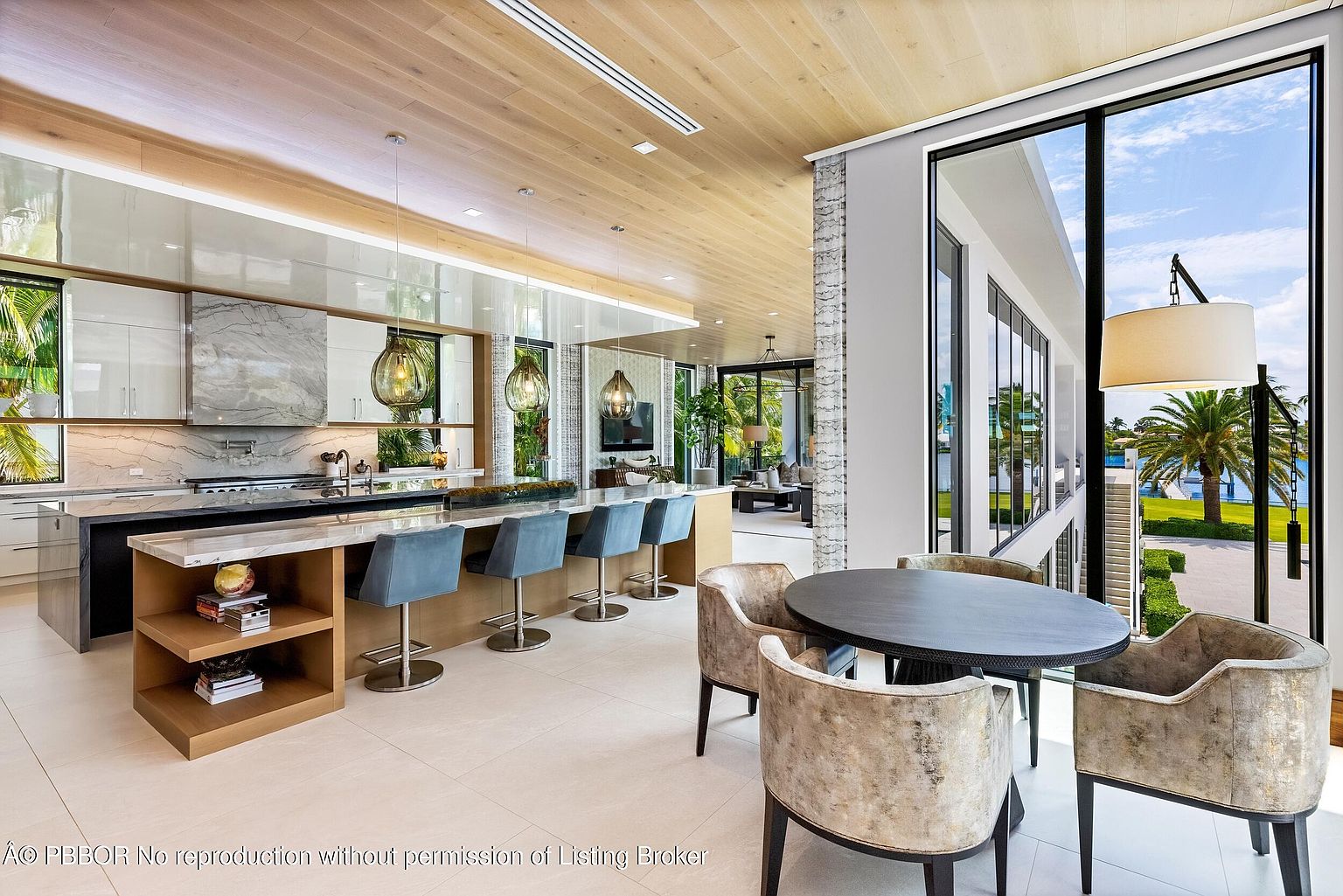 This is a bright and modern kitchen featuring a large island with seating, sleek cabinetry, and high-end appliances. The open floor plan seamlessly connects the kitchen to the dining area, which features a round table and stylish chairs, and the living area, creating an inviting space for entertaining. Large windows offer stunning views of the outdoors, enhancing the luxurious feel of the home.