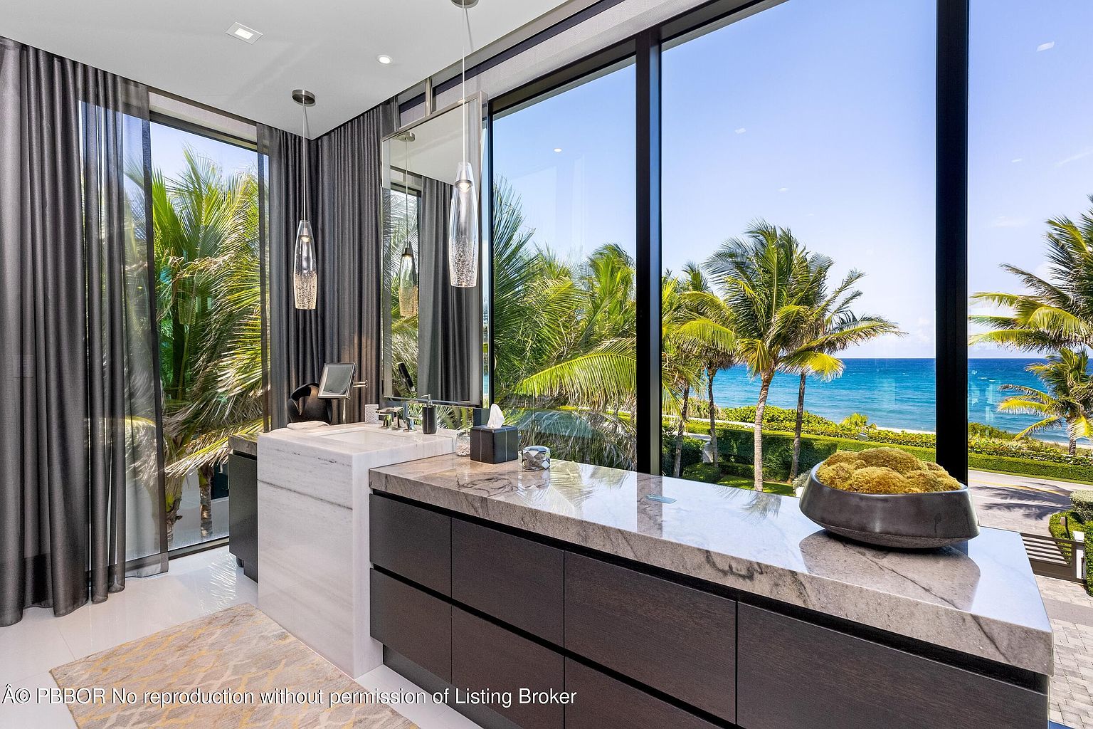 This is a luxurious primary bathroom featuring a modern vanity with a marble countertop and dark wood cabinetry. Large windows offer stunning views of palm trees and the ocean, creating a spa-like atmosphere. The room is well-lit with pendant lighting, and the overall design is sleek and sophisticated.