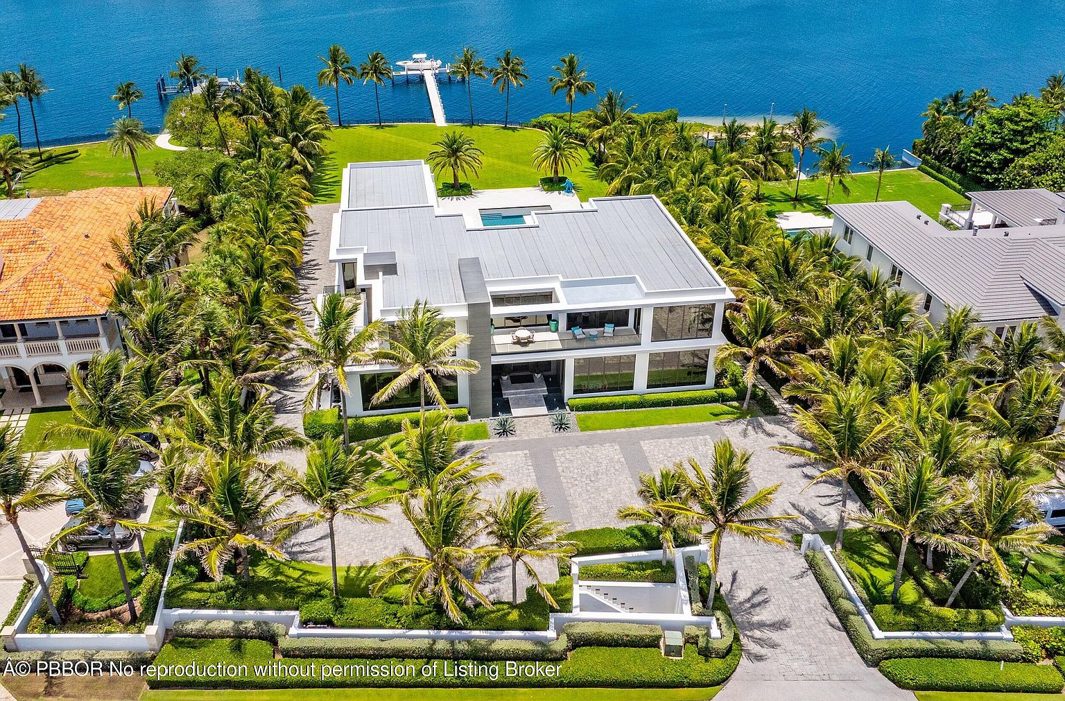 This aerial view showcases a modern, luxurious waterfront property. The house features a flat roof with a rooftop pool, large glass windows, and a meticulously landscaped yard with palm trees. A long dock extends into the blue water, adding to the property's appeal and exclusivity.