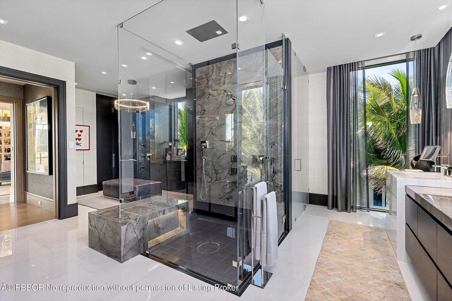 This is a luxurious primary bathroom featuring a glass-enclosed shower with marble walls and multiple shower heads. The bathroom has a modern design with sleek fixtures, a marble vanity, and large windows providing natural light and views of the exterior greenery. The space is bright and spacious, with a clean and elegant aesthetic.