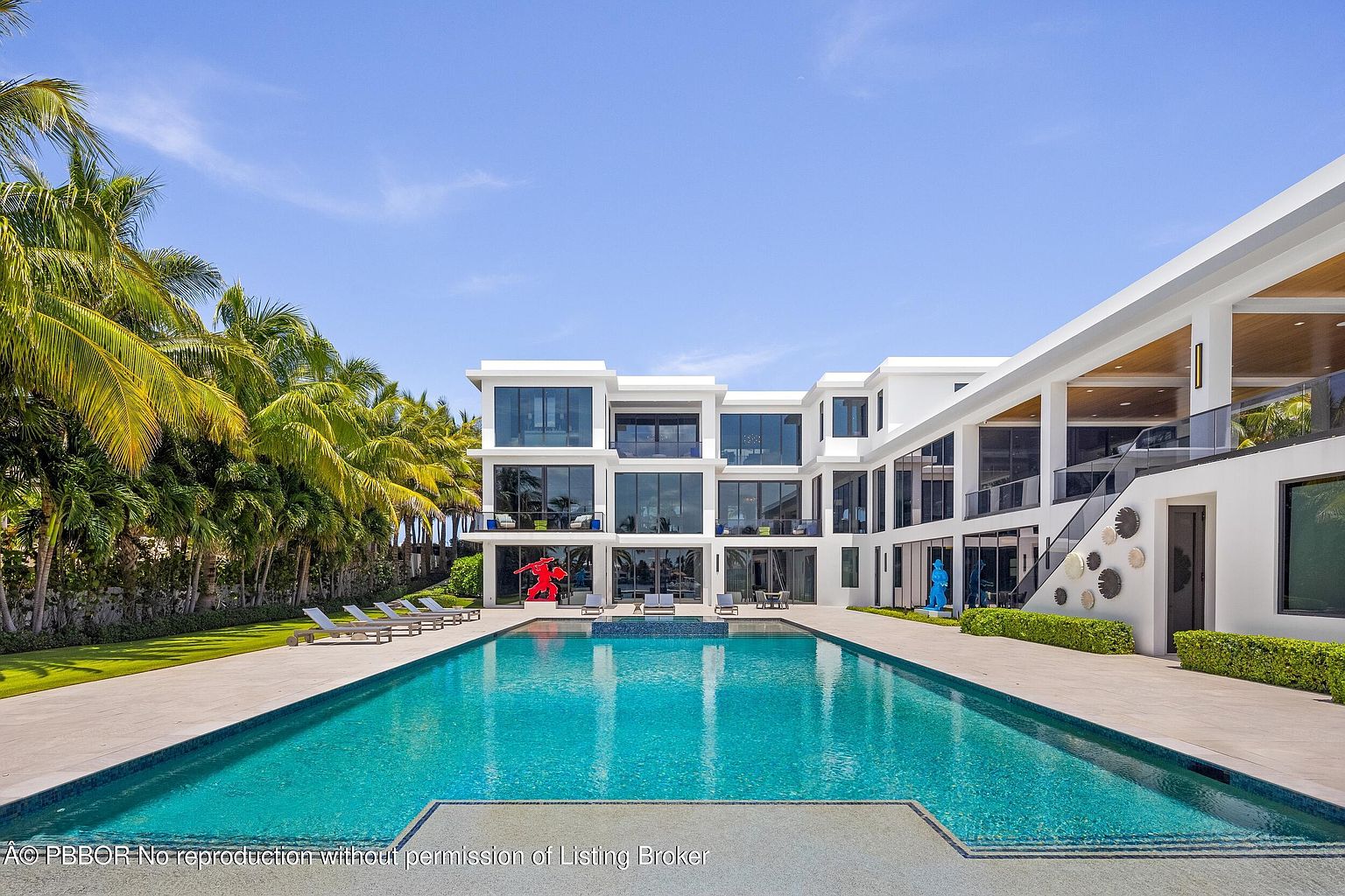 This image showcases the rear exterior of a modern luxury home, emphasizing a large rectangular swimming pool with clear blue water. The pool is surrounded by a spacious patio area with lounge chairs, and the house features clean architectural lines, large windows, and a multi-level design. Lush tropical landscaping adds to the property's appeal, creating a resort-like atmosphere.