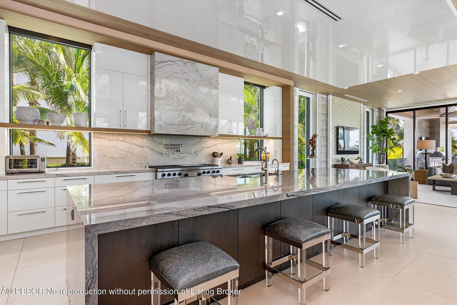 This is a bright and modern kitchen featuring a large island with seating, white cabinetry, and marble countertops and backsplash. The kitchen is well-lit with natural light from the windows, which offer a view of lush greenery. Stainless steel appliances and sleek fixtures add to the contemporary aesthetic.