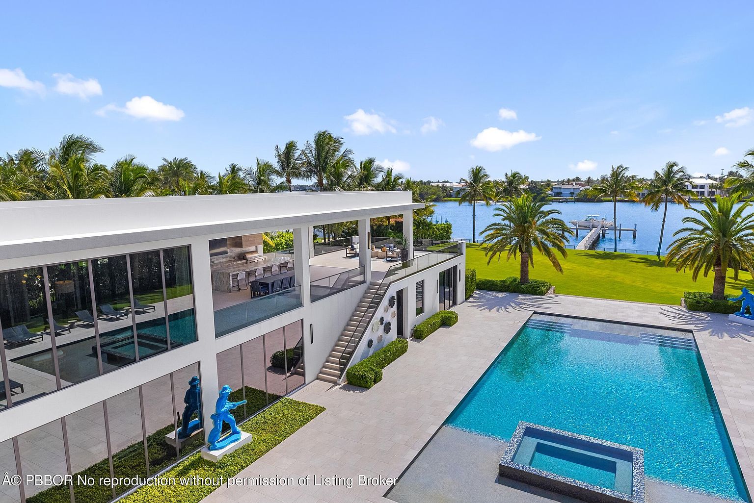 This aerial view showcases a luxurious modern home with a sleek, white exterior and expansive glass windows. The property features a large swimming pool with an integrated spa, a well-manicured lawn extending to the waterfront, and a spacious outdoor living area on the second floor. Palm trees and lush landscaping surround the property, enhancing its tropical appeal.