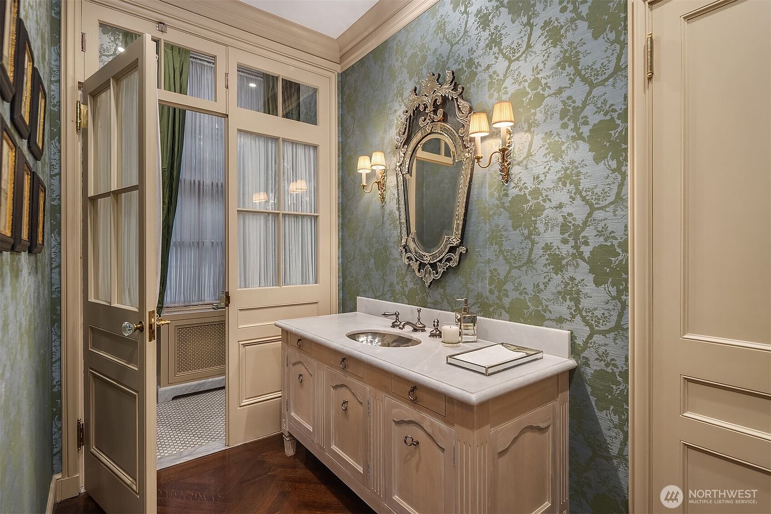 This elegant powder room features a sophisticated vanity with a marble countertop and an ornate, vintage-style mirror flanked by dual wall sconces. The walls are adorned with a classic green and blue botanical wallpaper, complemented by rich hardwood flooring and a set of French doors that open into an adjacent space. The overall aesthetic is refined and traditional, creating a luxurious and welcoming atmosphere.
