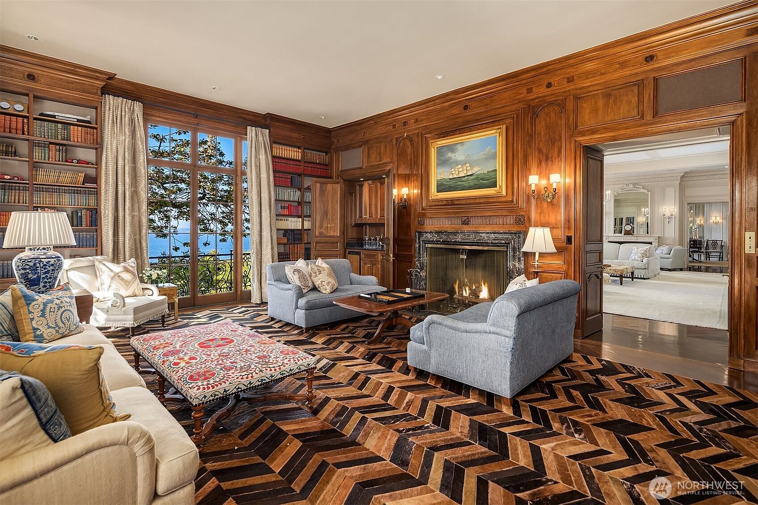This sophisticated wood-paneled study exudes classic elegance, featuring floor-to-ceiling built-in bookshelves, a grand fireplace with a marble surround, and a striking herringbone-patterned floor. The room is furnished with plush blue seating and a central ottoman, while large French doors provide a stunning view of the outdoors and allow for abundant natural light. The overall atmosphere is one of refined luxury and timeless comfort, perfect for a quiet retreat or professional workspace.