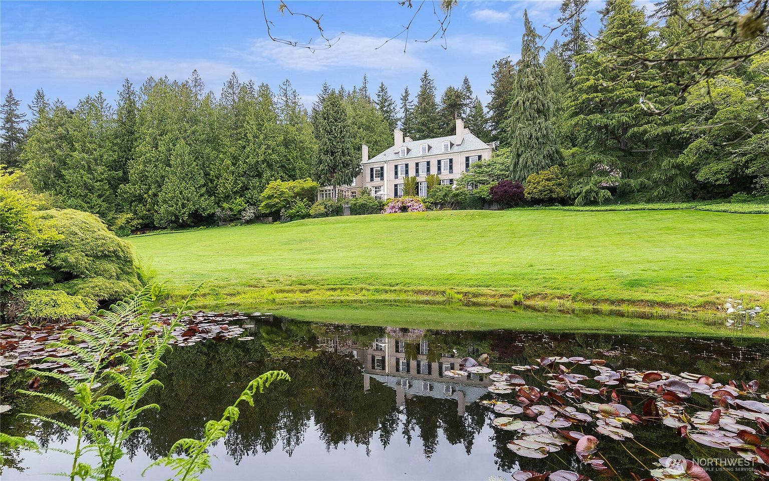 This picturesque estate features a grand, multi-story colonial-style mansion set against a backdrop of lush, mature evergreen trees. In the foreground, a tranquil pond filled with lily pads reflects the home's architecture, while a sprawling, manicured lawn gently slopes upward toward the residence. The scene evokes a sense of serene luxury and timeless elegance, perfectly capturing the expansive and private nature of the property.