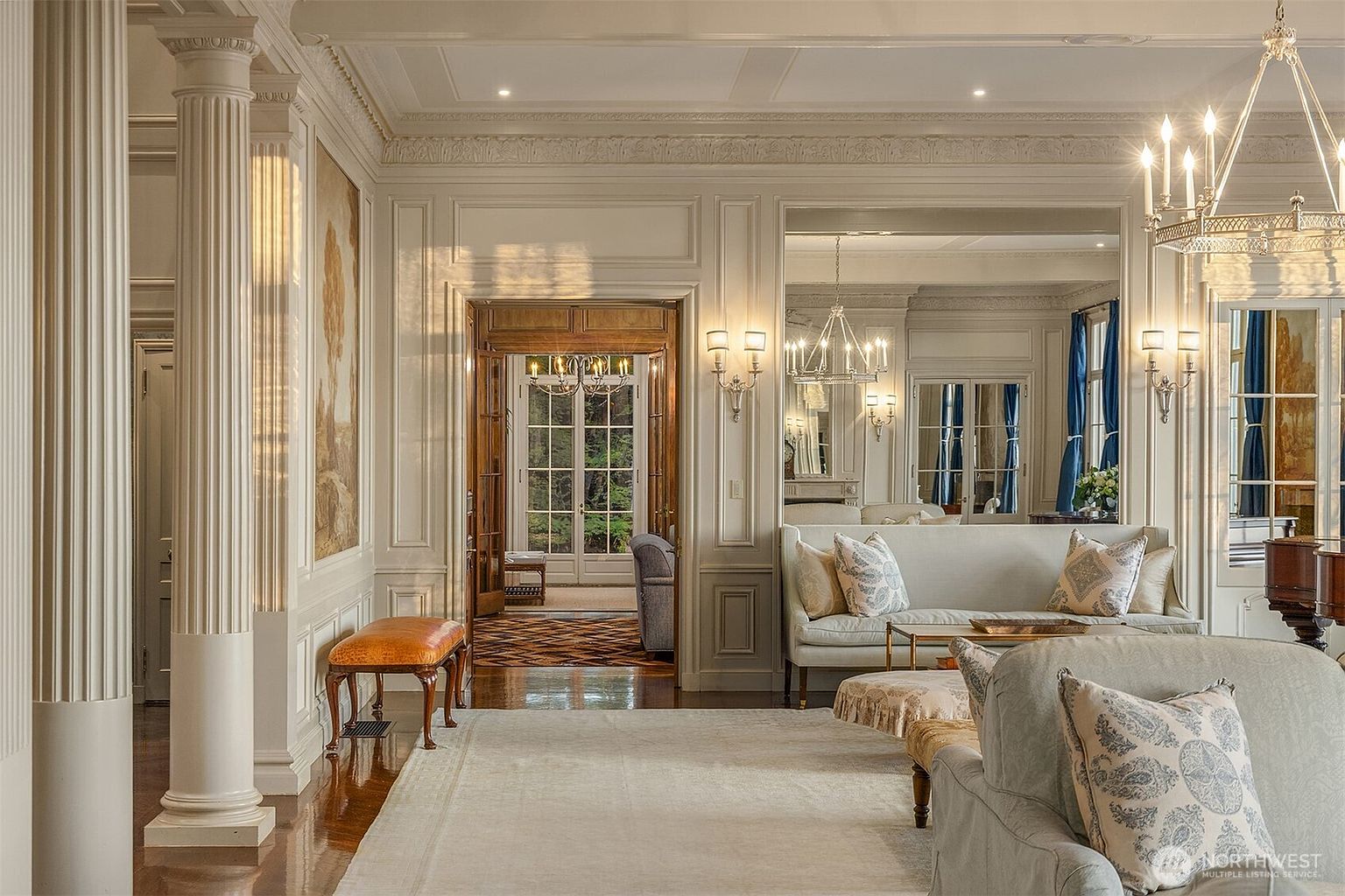 This elegant, formal living room exudes classic luxury with its high ceilings, intricate crown molding, and grand fluted columns. The space features a neutral color palette, plush seating, and a large area rug, all illuminated by a sophisticated crystal chandelier. A wide doorway leads to an adjacent room, creating a sense of depth and architectural grandeur that defines the home's refined aesthetic.