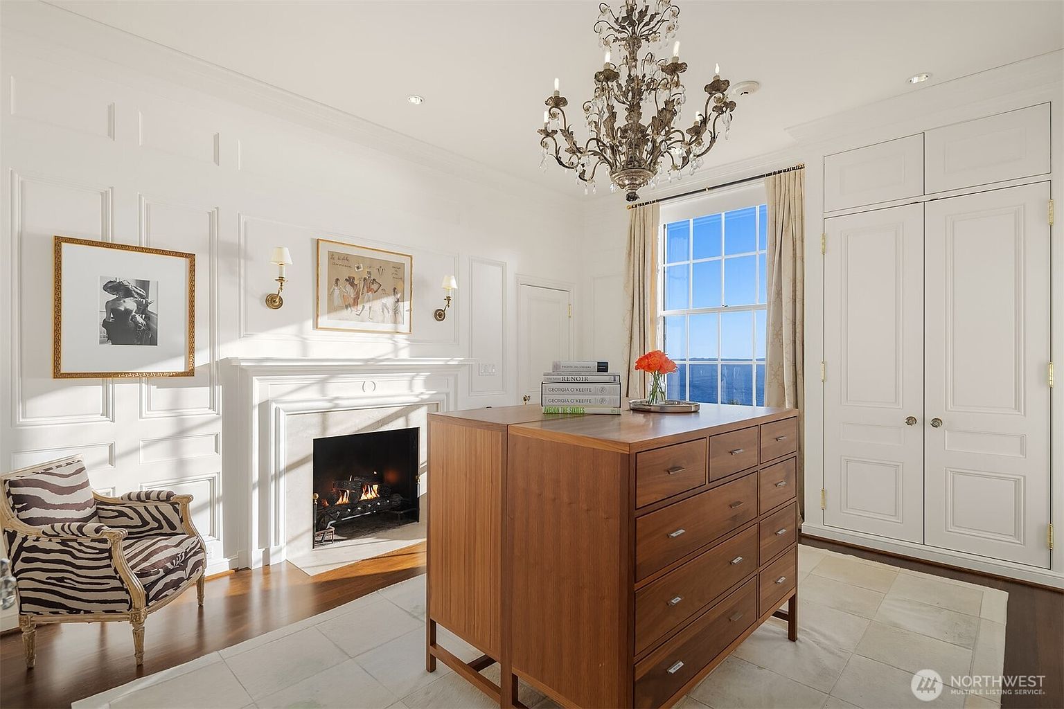 This elegant walk-in closet features a sophisticated design with white paneled walls, built-in cabinetry, and a central wooden dresser island. A classic crystal chandelier hangs above, while a zebra-print armchair and a fireplace add a touch of luxury and warmth to the space. The room is brightened by natural light from a large window that offers a scenic view, creating a refined and organized atmosphere.