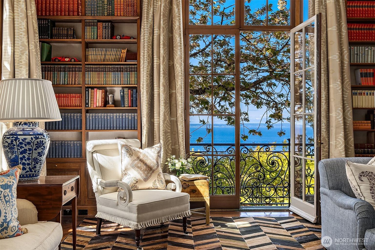 This elegant study features floor-to-ceiling wooden bookshelves filled with books and decorative model cars, creating a sophisticated and scholarly atmosphere. A plush armchair sits on a patterned rug, positioned perfectly to enjoy the stunning, unobstructed view of the ocean through the large French doors that open onto a wrought-iron balcony. The room exudes a classic, refined aesthetic, blending traditional library charm with a breathtaking coastal backdrop.