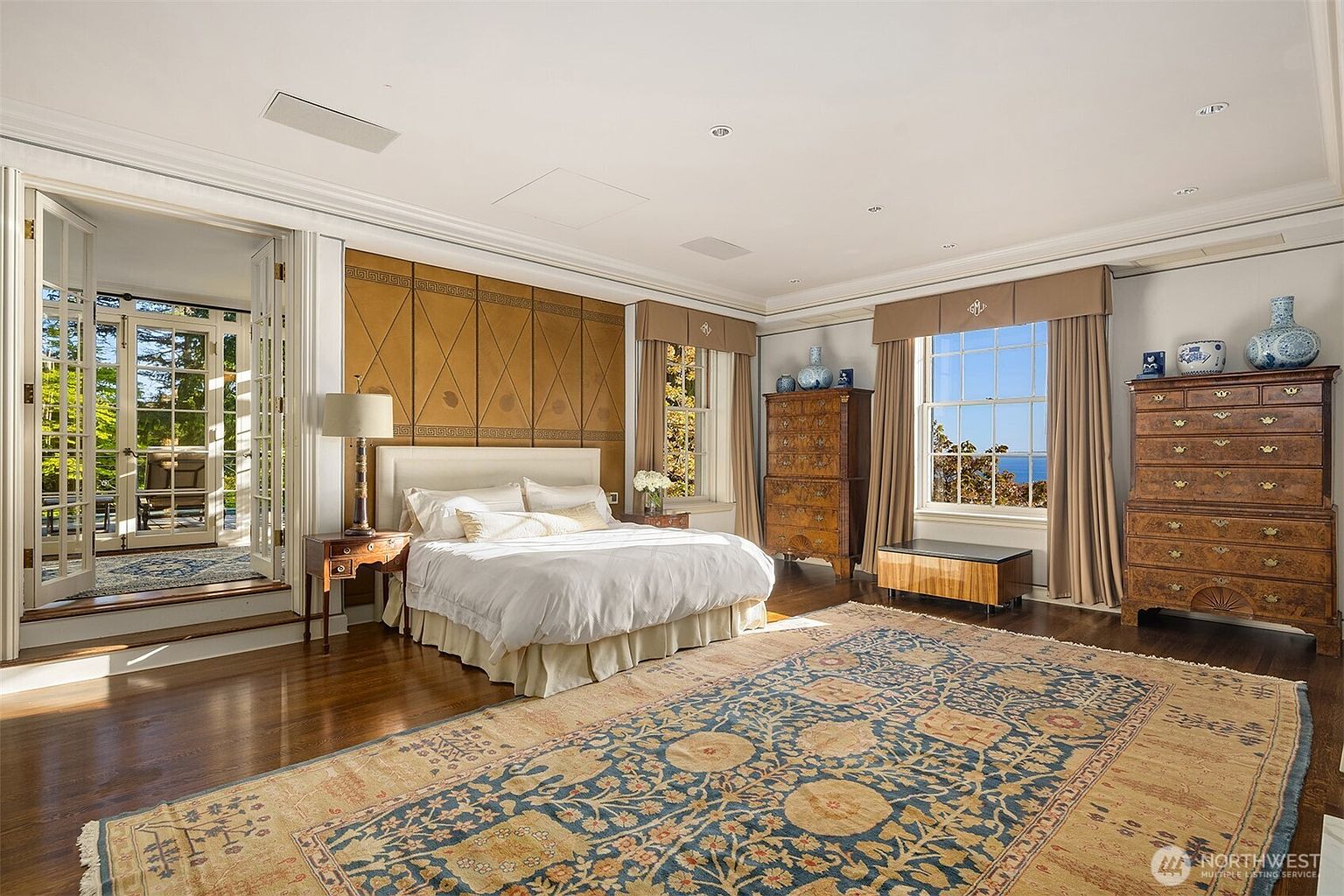 This spacious primary bedroom features a classic, elegant design with a large bed centered against a decorative wood-paneled wall. The room is bathed in natural light from large windows and French doors that open onto a private terrace, offering a serene view of the outdoors. Rich hardwood floors, a large patterned area rug, and antique-style wooden dressers contribute to the sophisticated and timeless atmosphere.