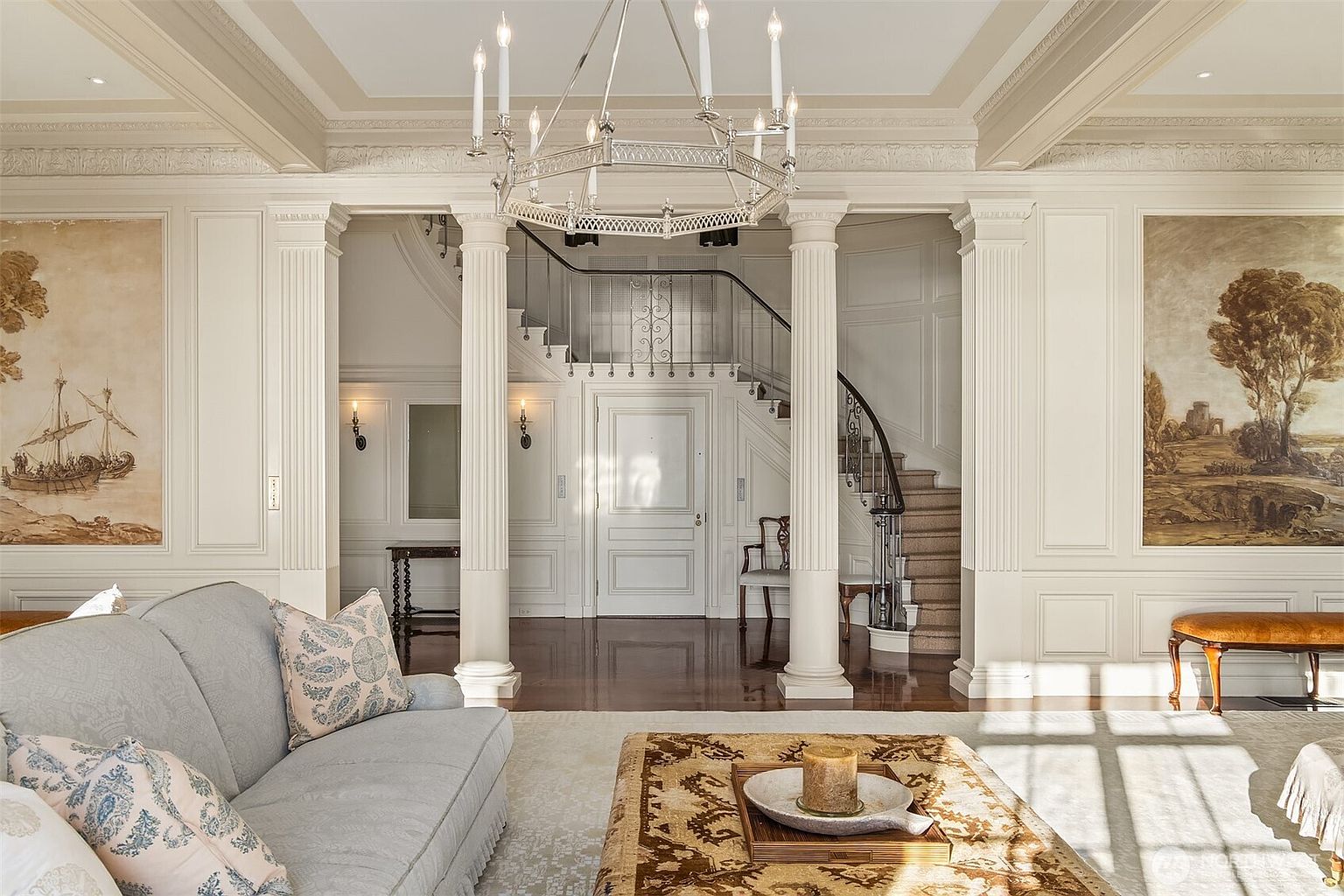 This elegant living room features a sophisticated, classic design with ornate wall molding, a grand chandelier, and two large framed landscape paintings. The space opens into a grand foyer with a sweeping staircase, framed by stately white columns that create a sense of architectural grandeur. The room is furnished with a plush light-colored sofa and a patterned ottoman, bathed in soft, natural light that highlights the polished hardwood floors.