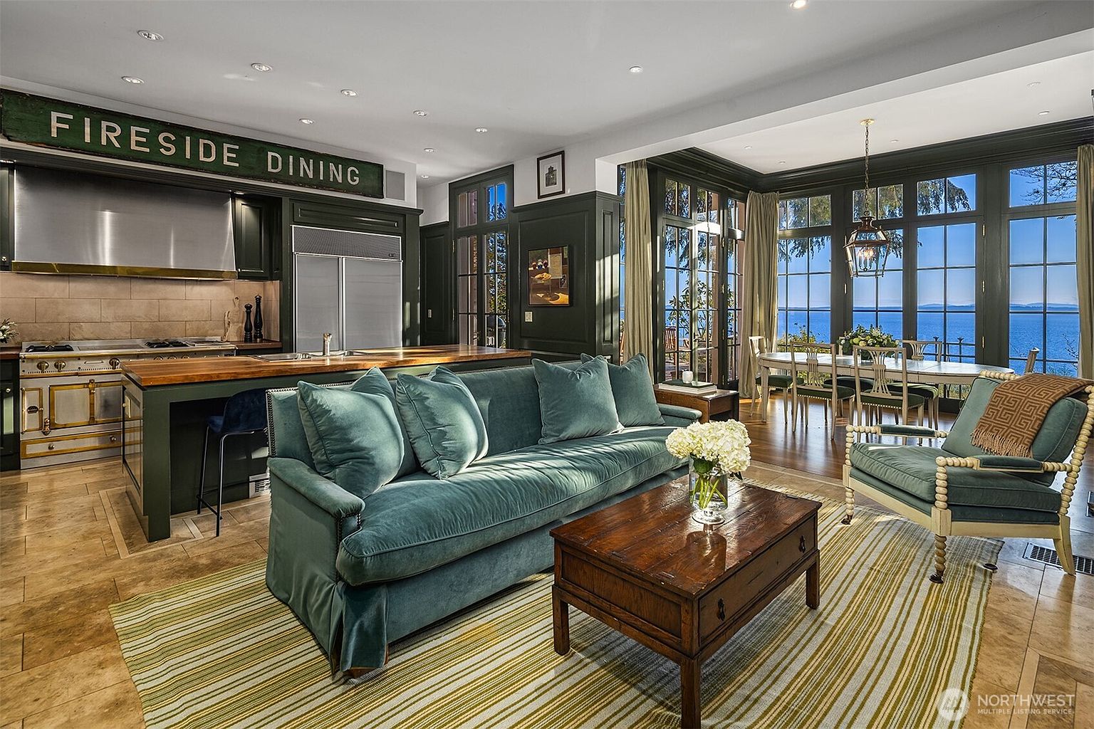 This elegant, open-concept living space seamlessly blends a cozy lounge area with a gourmet kitchen and a formal dining room overlooking a stunning waterfront view. The room features a plush teal velvet sofa, a rich wooden coffee table, and dark cabinetry that contrasts beautifully with the light-filled, expansive windows. The overall aesthetic is sophisticated and inviting, emphasizing a luxurious coastal lifestyle with high-end finishes and a warm, layered color palette.