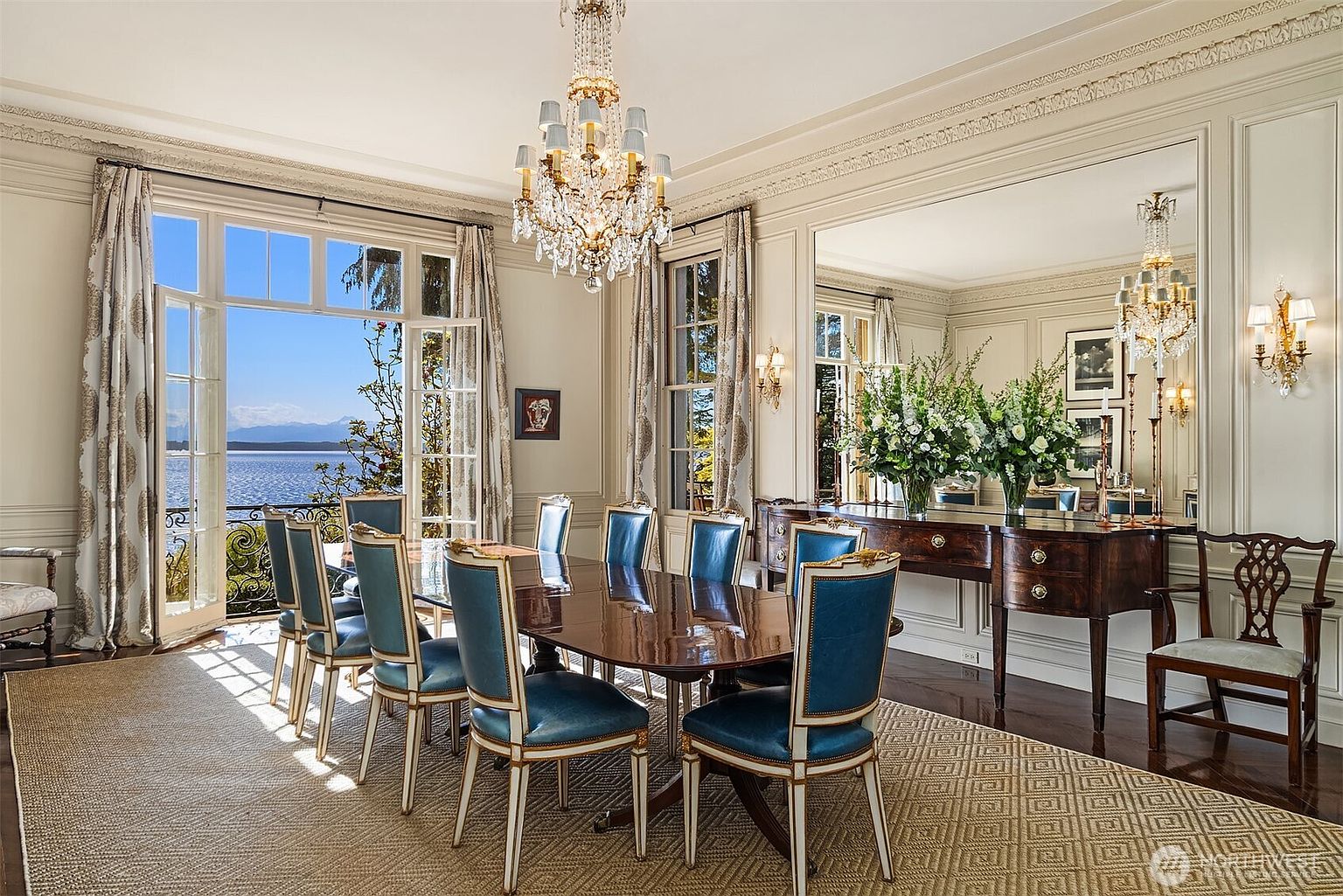 This elegant formal dining room features a polished mahogany table surrounded by blue upholstered chairs, set beneath a grand crystal chandelier. The room is framed by floor-to-ceiling windows that open onto a balcony with a stunning waterfront view, while a large wall mirror reflects the space and adds depth. The sophisticated decor, including crown molding and a patterned area rug, creates a luxurious and inviting atmosphere perfect for hosting.