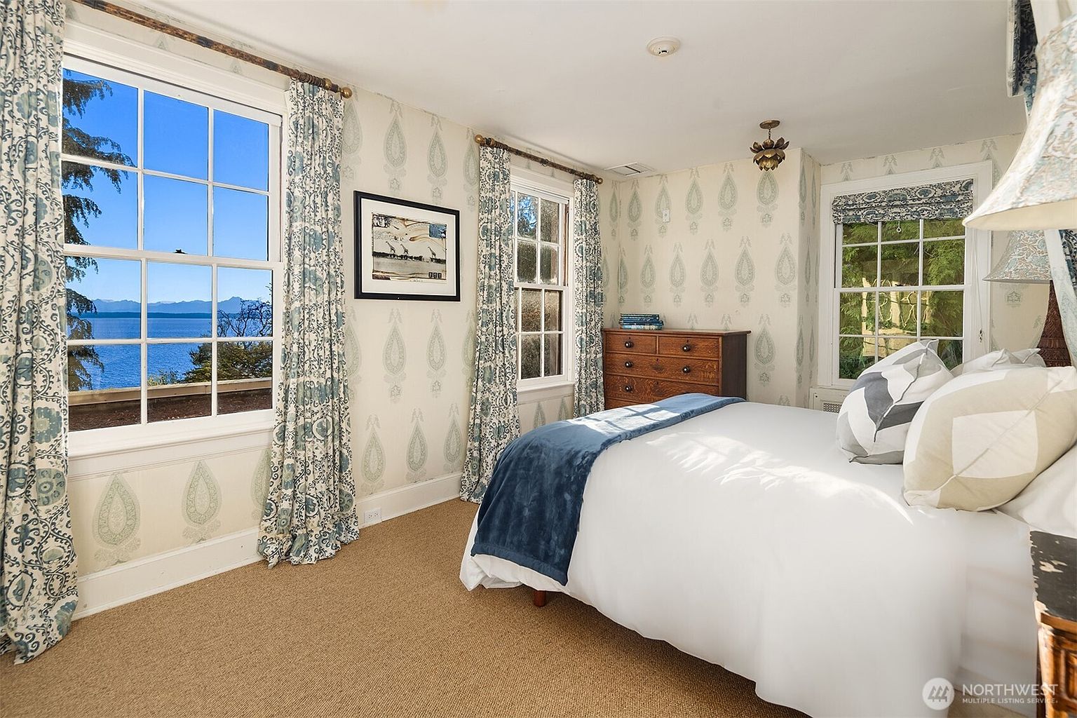This charming bedroom features a classic aesthetic with patterned wallpaper, elegant blue and white floral curtains, and a plush bed dressed in white linens with a navy throw. Large windows provide abundant natural light and a stunning, expansive view of the water and distant mountains. The room is anchored by a warm-toned wooden dresser, creating a cozy and inviting atmosphere.