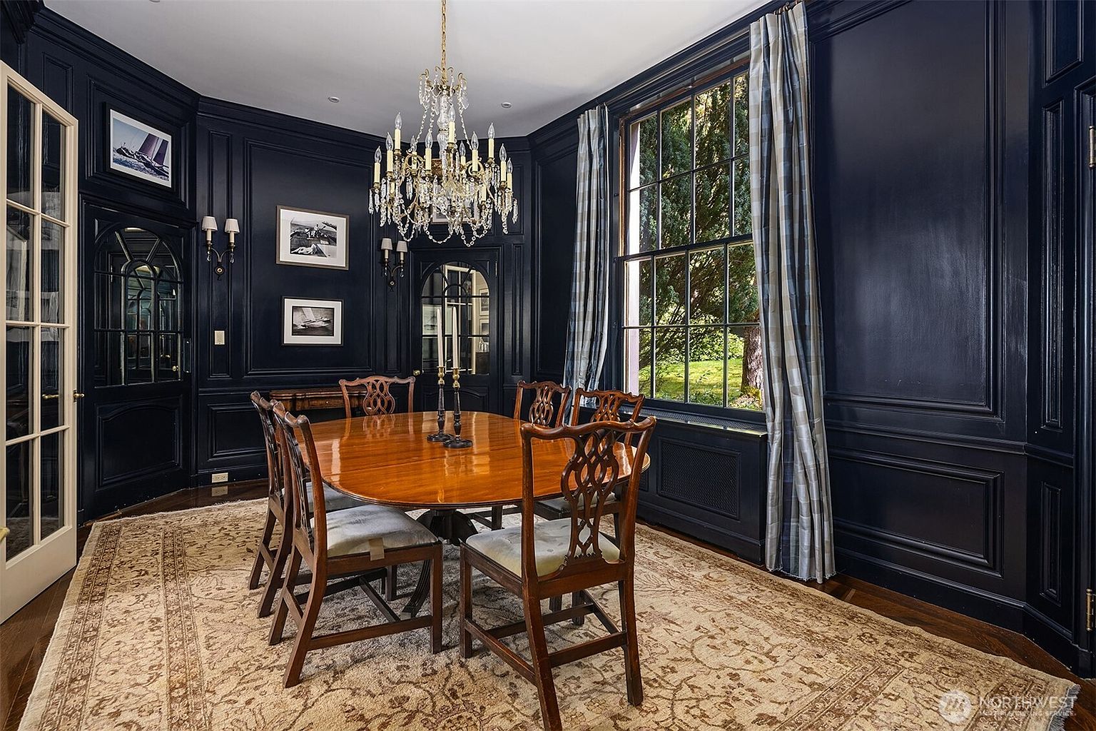 This elegant dining room features dramatic, high-gloss navy blue paneled walls that create a sophisticated and moody atmosphere. A large, polished wooden oval table sits at the center, surrounded by classic Chippendale-style chairs, all anchored by a large, intricate area rug. A grand crystal chandelier hangs above, while a tall window with floor-to-ceiling plaid curtains provides a view of the lush greenery outside.
