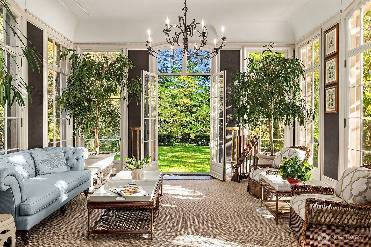 This elegant sunroom features a sophisticated blend of classic and natural elements, highlighted by a light blue tufted sofa and wicker armchairs arranged around a central coffee table. Large floor-to-ceiling windows and French doors flood the space with natural light, offering a seamless transition to the lush green garden outside. The room is accented by potted trees, a decorative wrought-iron chandelier, and framed botanical art, creating a serene and inviting atmosphere.