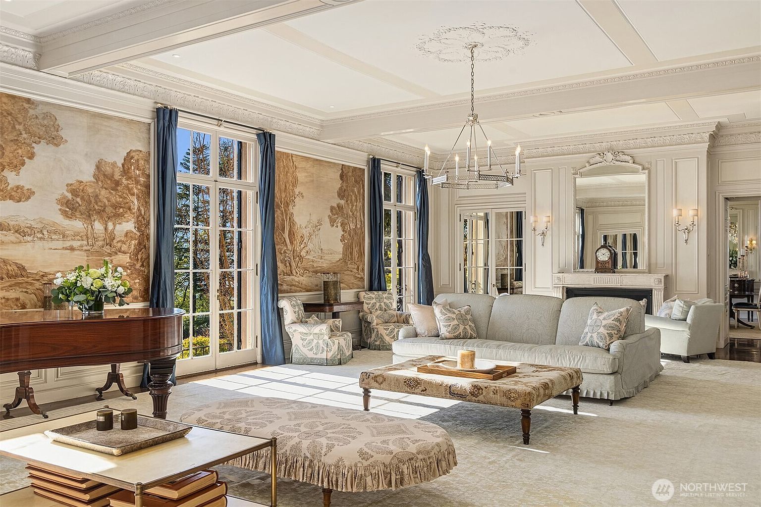 This elegant, formal living room exudes classic sophistication with its high coffered ceilings, intricate crown molding, and expansive floor-to-ceiling windows draped in deep blue curtains. The space is anchored by a plush, light-toned sofa and a patterned ottoman, complemented by a grand piano and scenic mural wallpaper that adds a touch of old-world charm. The perspective is a wide-angle shot from the corner of the room, capturing the luxurious furnishings and the bright, airy atmosphere.