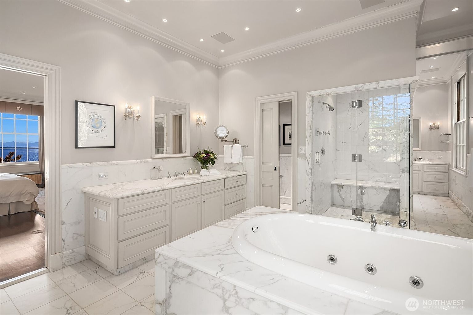 This luxurious primary bathroom features a spacious marble-clad soaking tub as the centerpiece, complemented by a double vanity with marble countertops and a large walk-in glass shower. The room is bathed in bright, neutral tones, creating an elegant and serene atmosphere, with a glimpse into an adjacent bedroom offering a scenic water view. The high ceilings and recessed lighting enhance the sophisticated, spa-like aesthetic of the space.