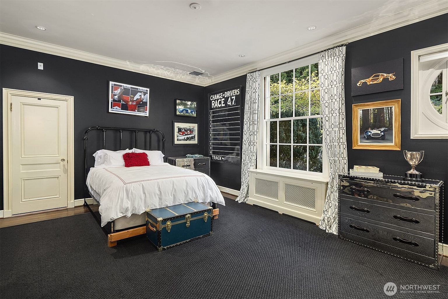 This themed bedroom features a bold, dark charcoal wall color contrasted by a white bed frame and bedding, creating a sophisticated yet playful racing-inspired aesthetic. The room is decorated with automotive-themed artwork, a custom race-track scoreboard, and a vintage-style trunk at the foot of the bed, all set against a large window with patterned curtains. The space is well-lit with recessed lighting and includes a sleek, dark dresser, offering a unique and stylish retreat.