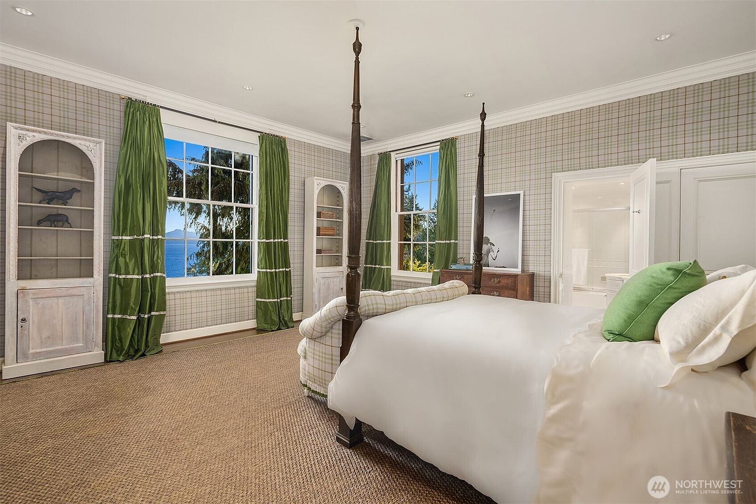 This elegant bedroom features a classic four-poster bed as the centerpiece, complemented by plaid wallpaper and vibrant green curtains that frame windows overlooking a scenic water view. The room includes two tall, arched-top wooden bookcases and a warm, textured carpet, creating a cozy and traditional atmosphere. The perspective is from the foot of the bed, looking toward the windows and an open doorway leading to an en-suite bathroom.