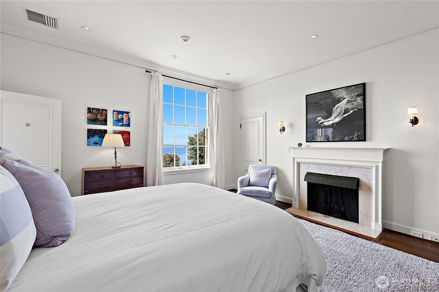 This elegant primary bedroom features a large, plush bed in the foreground and a classic fireplace with a mantelpiece displaying a striking black-and-white underwater photograph. A large window provides a serene view of the water, while a cozy armchair sits in the corner, creating a peaceful and sophisticated atmosphere. The room is finished with crisp white walls, dark hardwood floors, and a soft area rug, emphasizing a bright and airy aesthetic.