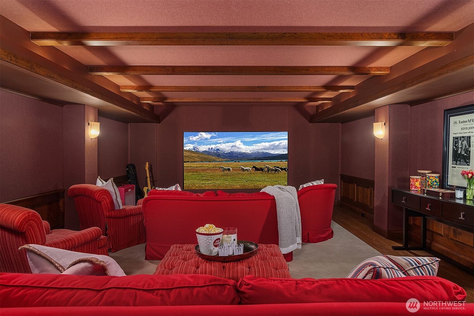 This cozy, dedicated home theater room features a deep red color palette, plush red seating, and a large screen displaying a scenic landscape. The space is accented by warm wood ceiling beams, wall sconces, and a framed Elvis Presley poster, creating an inviting and cinematic atmosphere. The perspective is from the back of the room, looking toward the screen, emphasizing the comfortable and immersive layout.