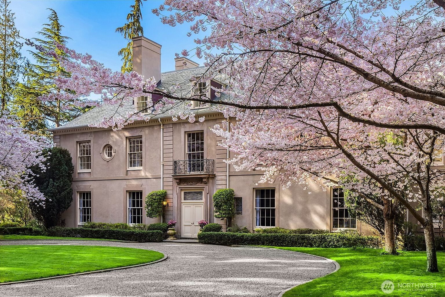 This elegant, multi-story estate features a classic architectural design with a light-colored stucco facade, symmetrical window placement, and a prominent central entryway with a decorative balcony above. The home is framed by lush, blooming cherry blossom trees, creating a picturesque and serene spring atmosphere. A manicured lawn and a curved gravel driveway lead up to the front entrance, emphasizing the property's grand curb appeal.