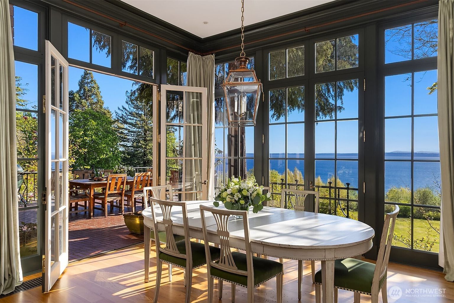 This elegant dining room features a light-colored oval table surrounded by classic chairs with green upholstered seats, set against a backdrop of floor-to-ceiling windows that offer a stunning panoramic view of the ocean. The room is framed by dark-painted window casings and a sophisticated lantern-style pendant light, creating a seamless transition to the adjacent outdoor brick patio. The overall atmosphere is bright, airy, and luxurious, emphasizing the home's connection to its scenic waterfront location.