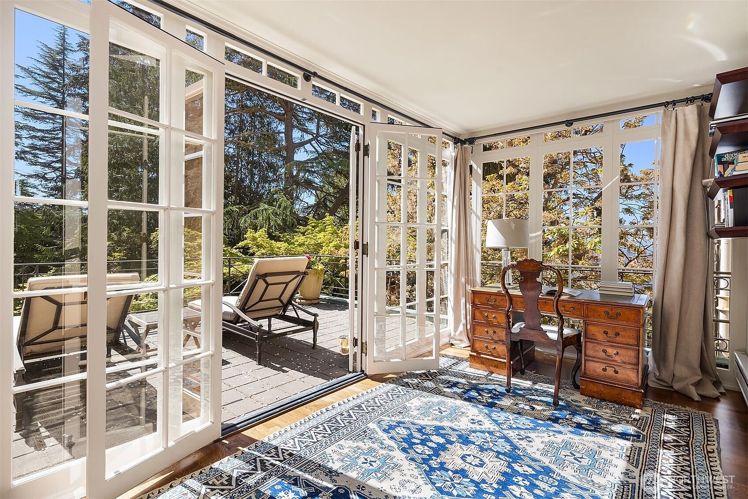 This elegant home office features a classic wooden desk and a traditional chair, set against a backdrop of expansive floor-to-ceiling French doors that open onto a sun-drenched deck. The room is anchored by a large, intricate blue and white patterned area rug, while the surrounding windows offer a serene, tree-filled view. The space exudes a sophisticated, tranquil atmosphere, perfect for focused work while enjoying the connection to the outdoors.