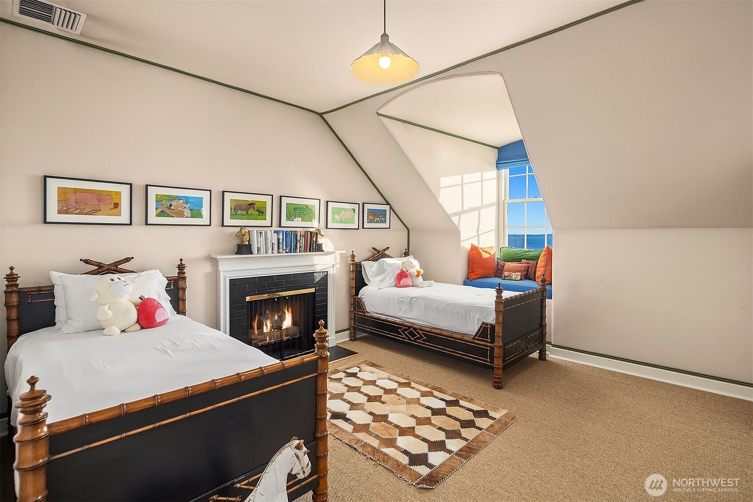 This charming attic-style bedroom features two twin beds with dark bamboo-style frames, positioned on either side of a cozy fireplace. The room is accented with a series of framed animal prints, a geometric patterned rug, and a built-in window seat that offers a scenic view of the water. The slanted ceilings and soft neutral walls create a warm, inviting atmosphere perfect for a guest room or children's space.