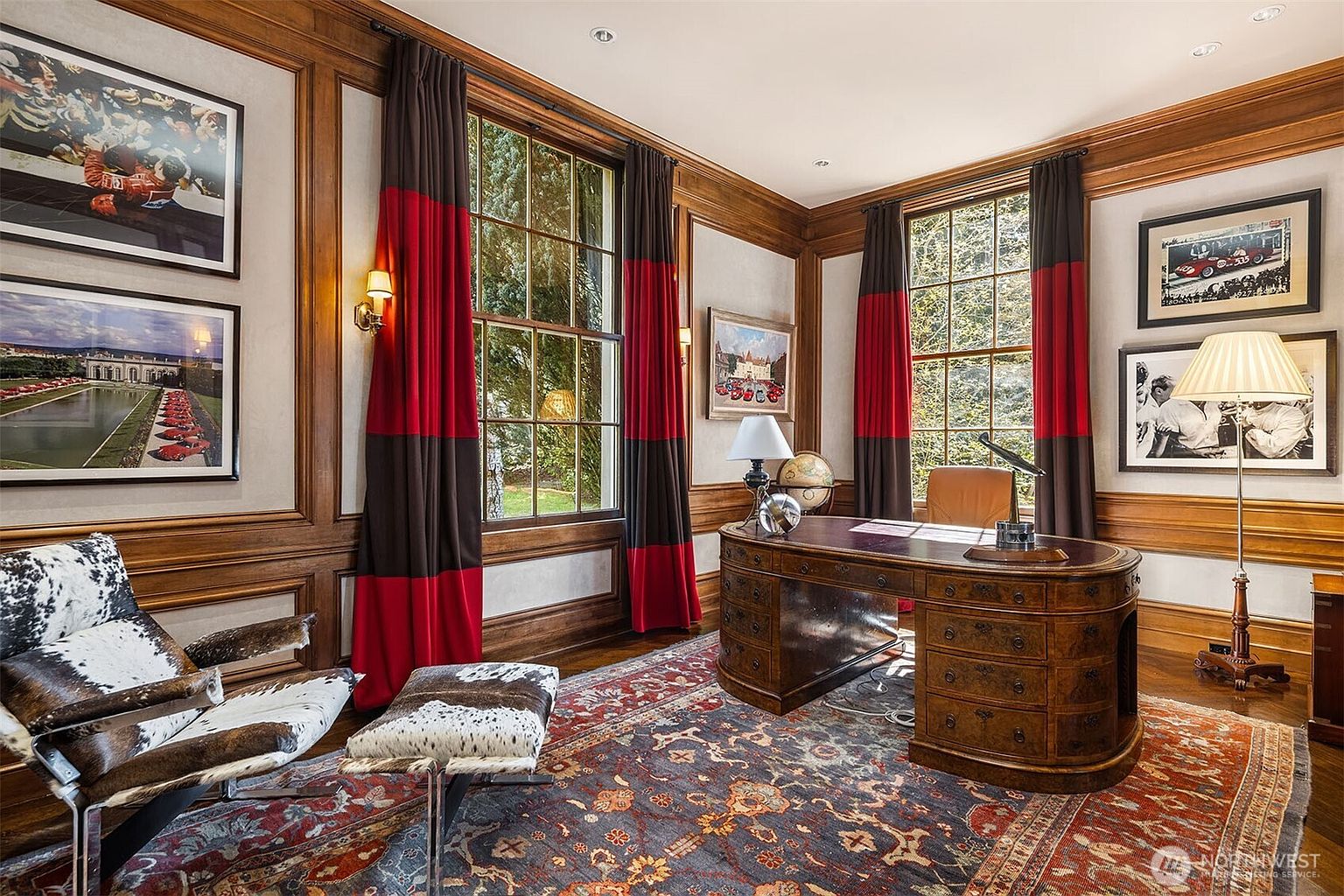 This sophisticated home office features rich wood-paneled walls, a large antique-style executive desk, and a patterned area rug that anchors the space. The room is accented with cowhide-upholstered seating, framed automotive-themed artwork, and floor-to-ceiling windows draped in bold red and brown curtains. The overall atmosphere is one of classic, masculine elegance, perfect for a professional study or library.