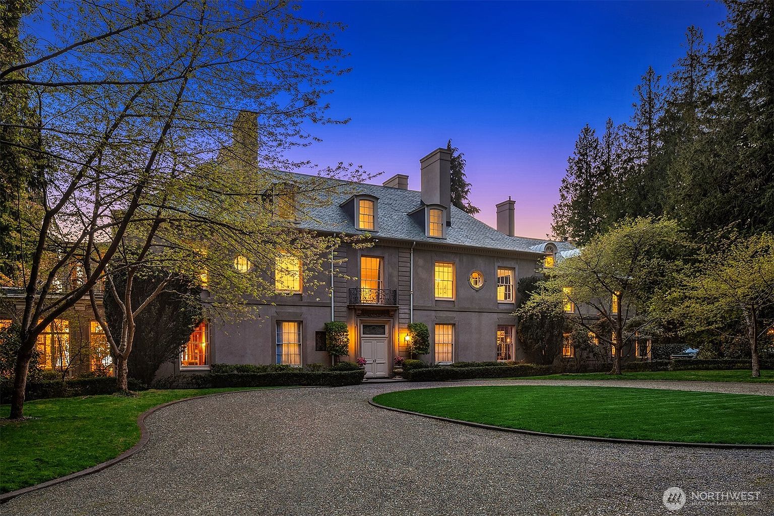 This grand, multi-story estate features a classic architectural style with a symmetrical facade, steep rooflines, and numerous illuminated windows that create a warm, inviting glow against the twilight sky. A circular gravel driveway leads up to the central entryway, framed by manicured hedges and mature trees that enhance the property's stately curb appeal. The perspective is a wide, eye-level shot that captures the full scale and elegance of the residence in a serene, dusk setting.