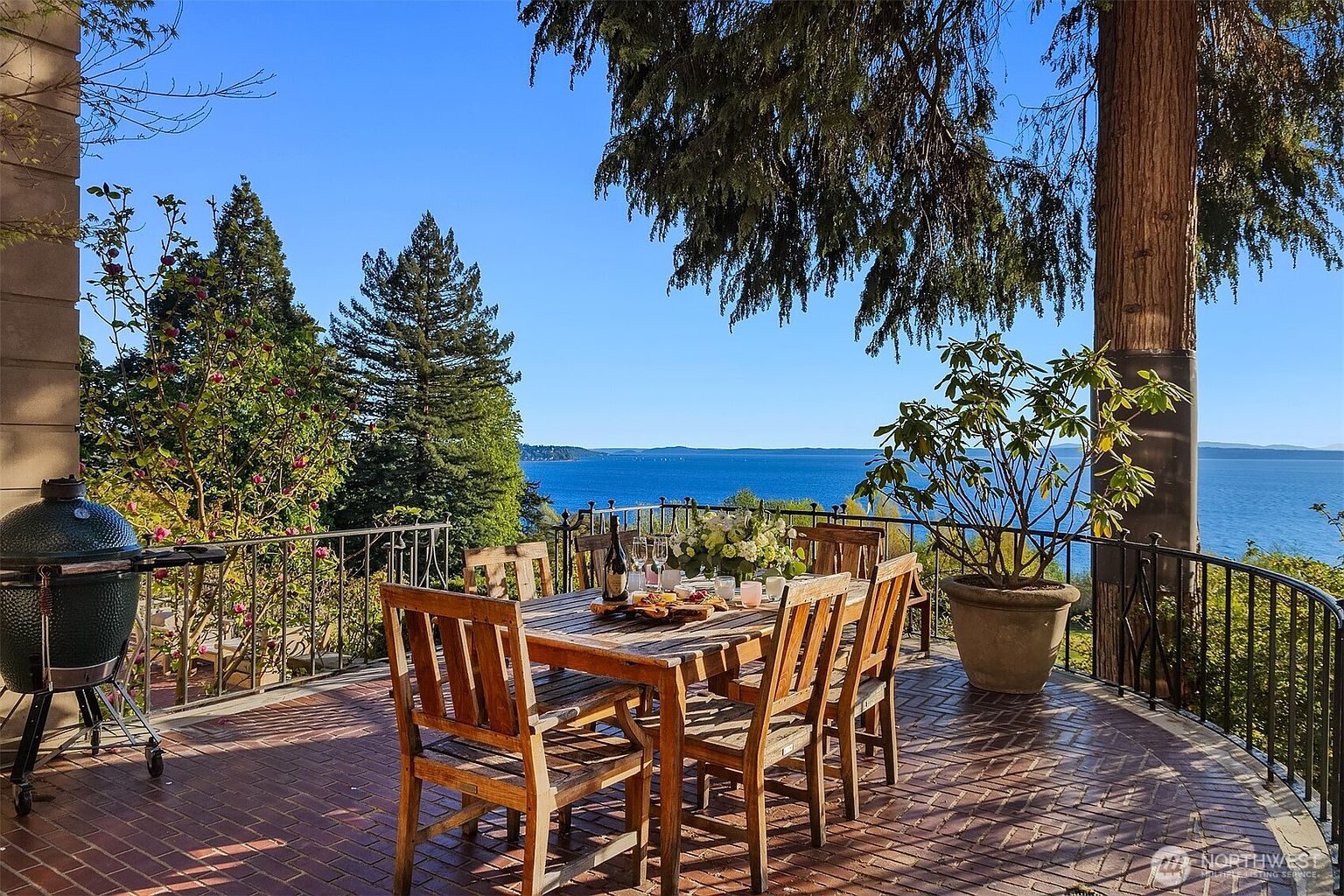 This inviting outdoor patio features a brick-paved floor and a wooden dining set arranged for an elegant meal overlooking a stunning, expansive water view. The space is framed by lush greenery, a large potted plant, and a classic wrought-iron railing, creating a serene and sophisticated atmosphere perfect for entertaining. The bright, clear sky and panoramic coastal scenery provide a picturesque backdrop that highlights the property's premium location.