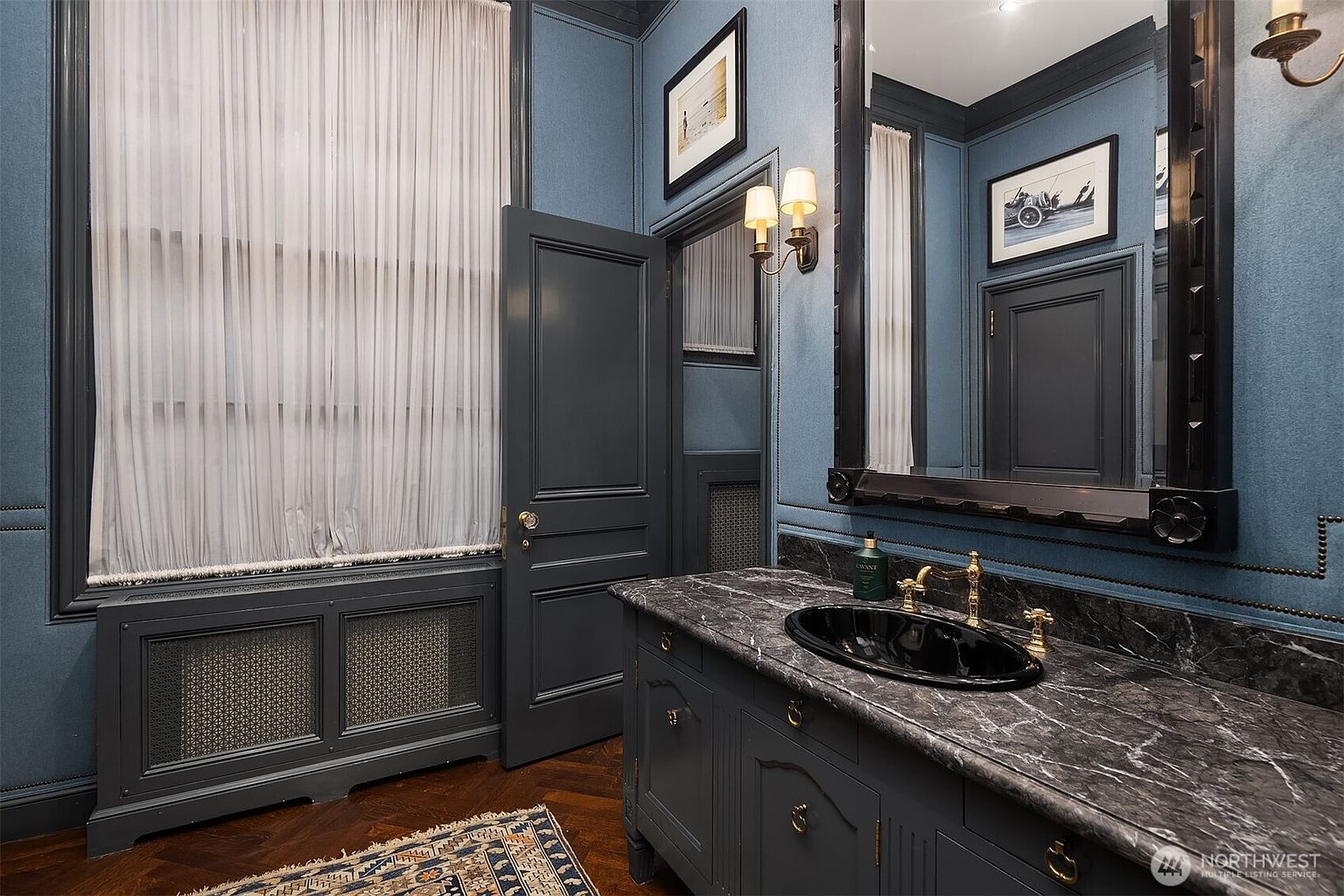 This sophisticated powder room features deep blue textured walls, dark wood trim, and a striking black marble vanity with a matching oval sink. The space is accented by elegant gold-toned hardware, a large framed mirror, and a classic herringbone hardwood floor, creating a moody and luxurious atmosphere. The perspective is a medium shot capturing the vanity and the architectural details of the room.