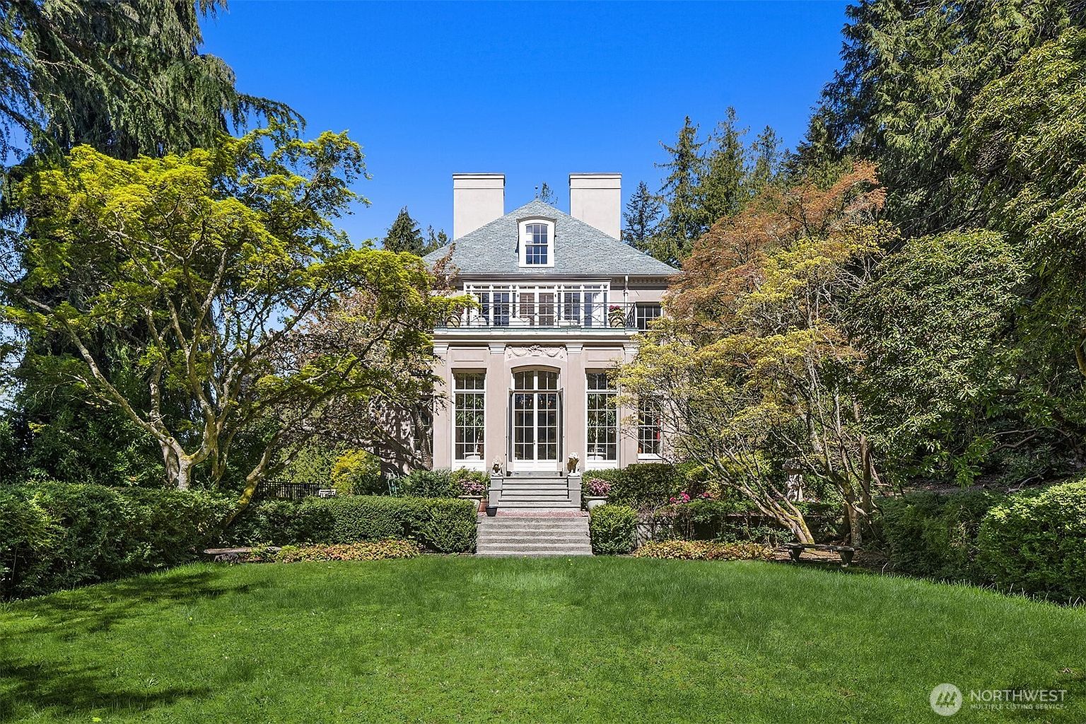 This elegant, multi-story estate features a classic architectural design with a symmetrical facade, tall arched windows, and a prominent second-floor balcony. The home is set against a backdrop of lush, mature trees and is framed by a meticulously maintained lawn and manicured garden beds. A stone staircase leads directly from the verdant backyard up to the main entrance, creating a grand and inviting transition between the outdoor landscape and the residence.