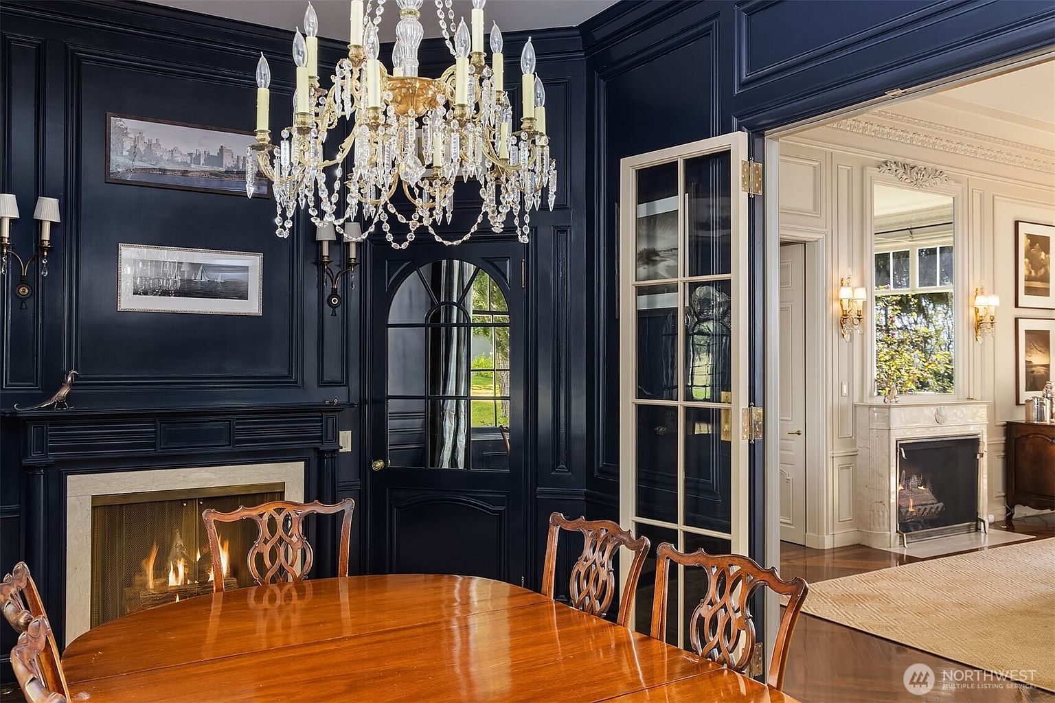 This elegant dining room features dramatic navy blue paneled walls, a grand crystal chandelier, and a polished wooden table, creating a sophisticated and formal atmosphere. A fireplace with a stone surround adds warmth, while glass-paned doors provide a view into an adjacent, lighter-toned room with its own fireplace. The perspective is from a low angle at the dining table, emphasizing the luxurious textures and architectural depth of the space.